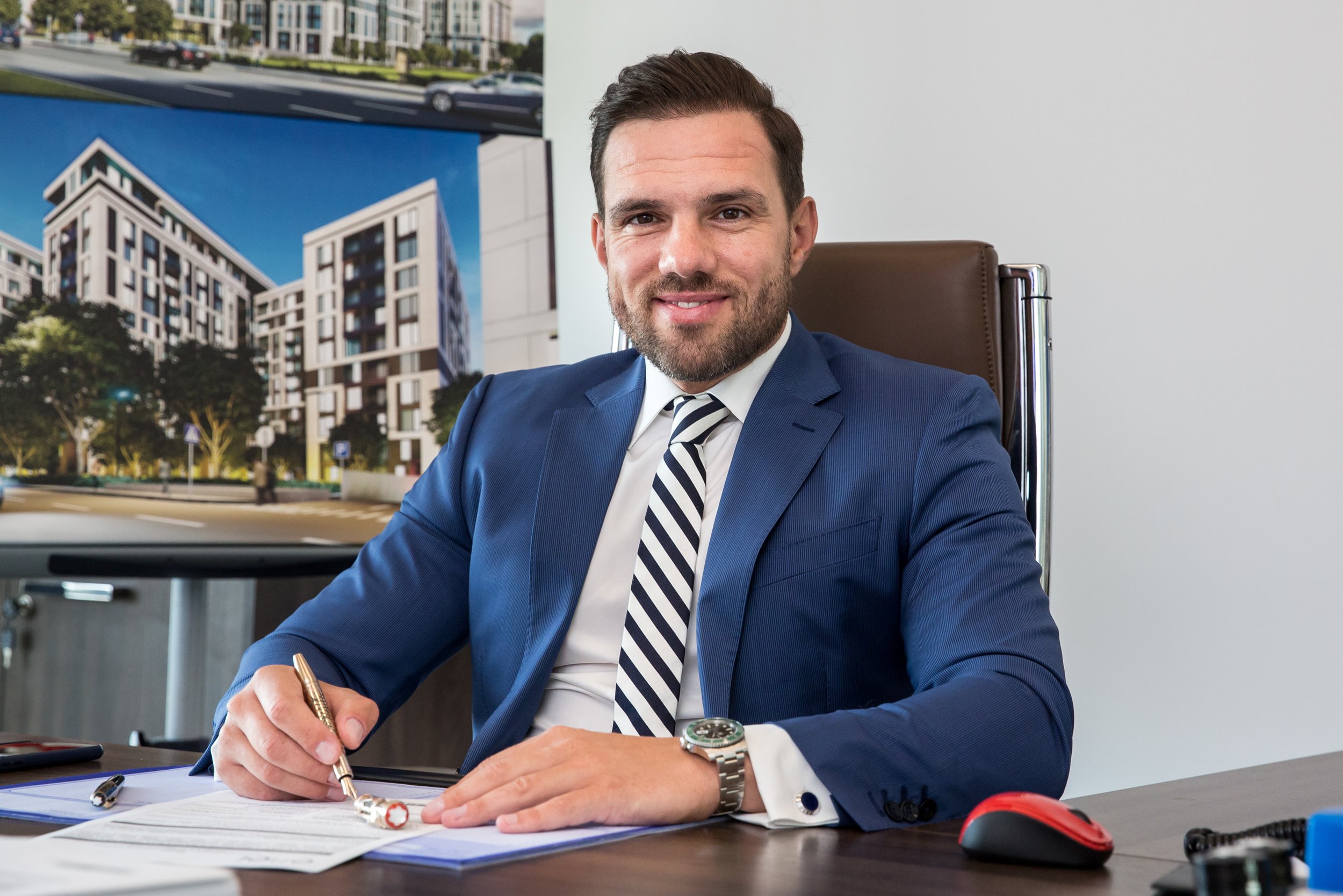 A man in a blue suit with a striped tie is sitting at a desk, smiling at the camera, holding a pen with papers in front of him. He has a wristwatch and is in an office with a white wall and architectural renderings of residential buildings on the wal