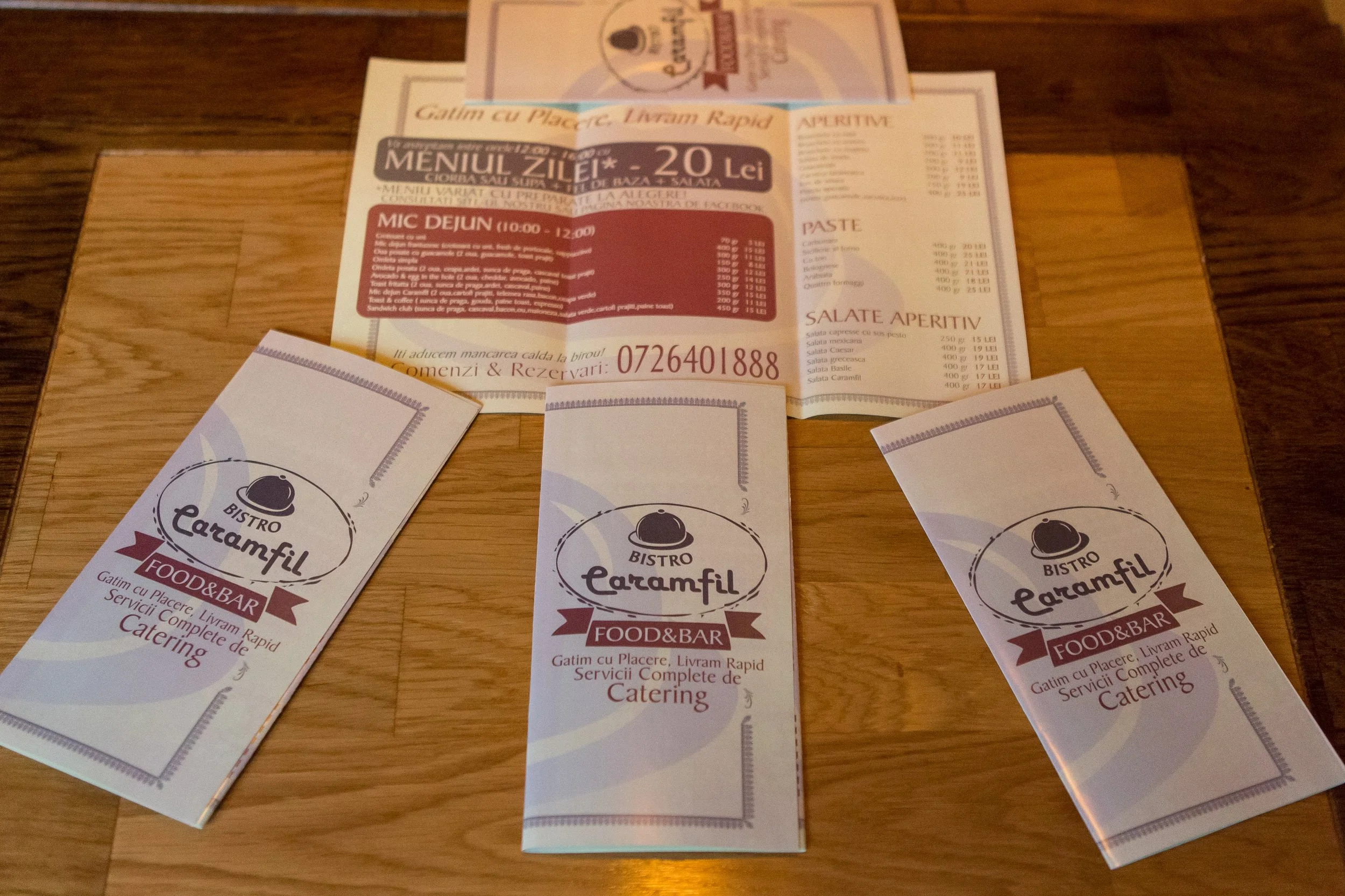 A collection of printed restaurant menus and business cards on a wooden table. The menus and cards are from Bistro Faramfil, a food and bar restaurant, and are written in Romanian. The menus and cards feature the restaurant's logo with a chef's hat a