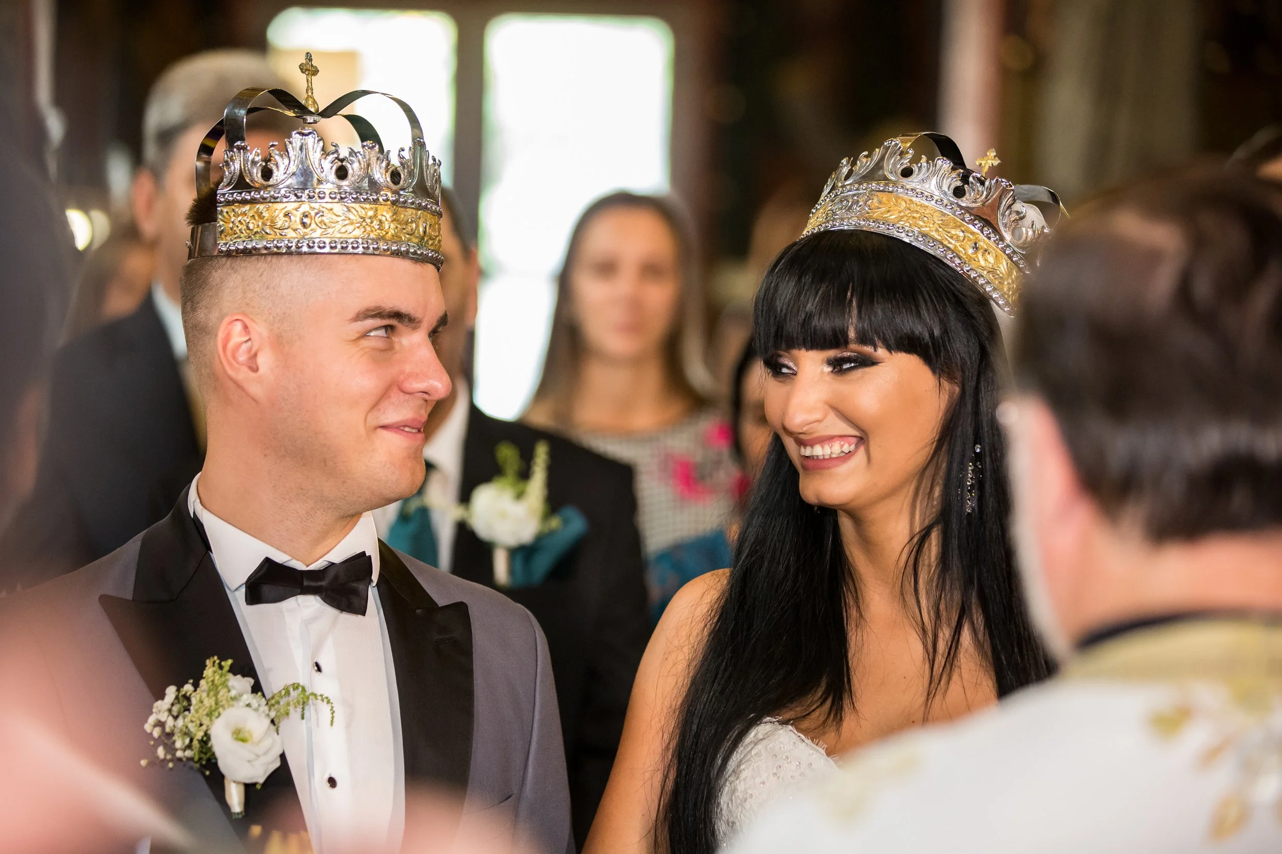 A couple during their wedding ceremony, both wearing crowns. The groom is in a tuxedo and the bride in a wedding dress, smiling at each other, surrounded by guests.
