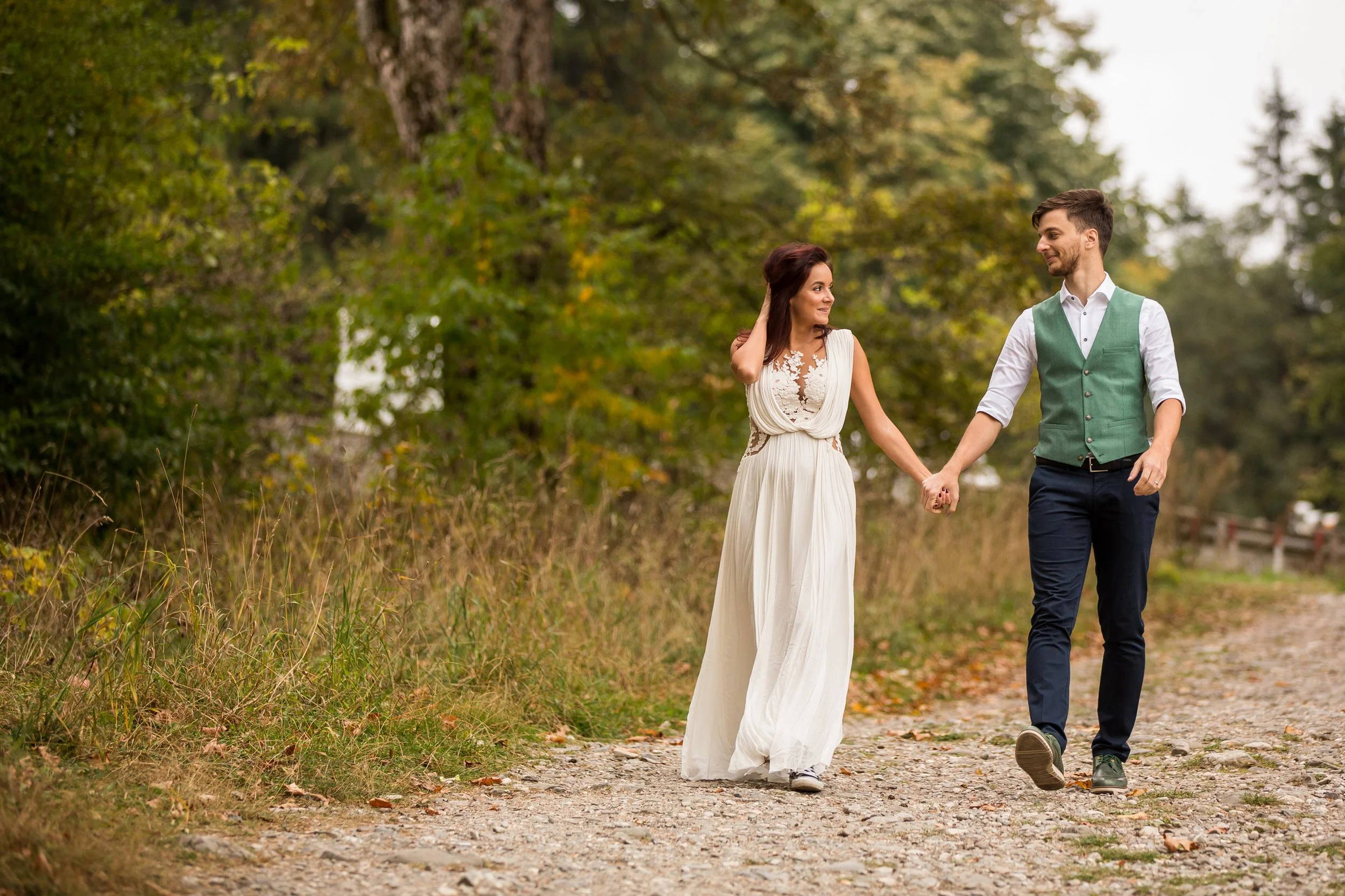 A couple walking hand in hand on a nature trail surrounded by trees, with the woman in a white dress and the man in a white shirt and green vest.