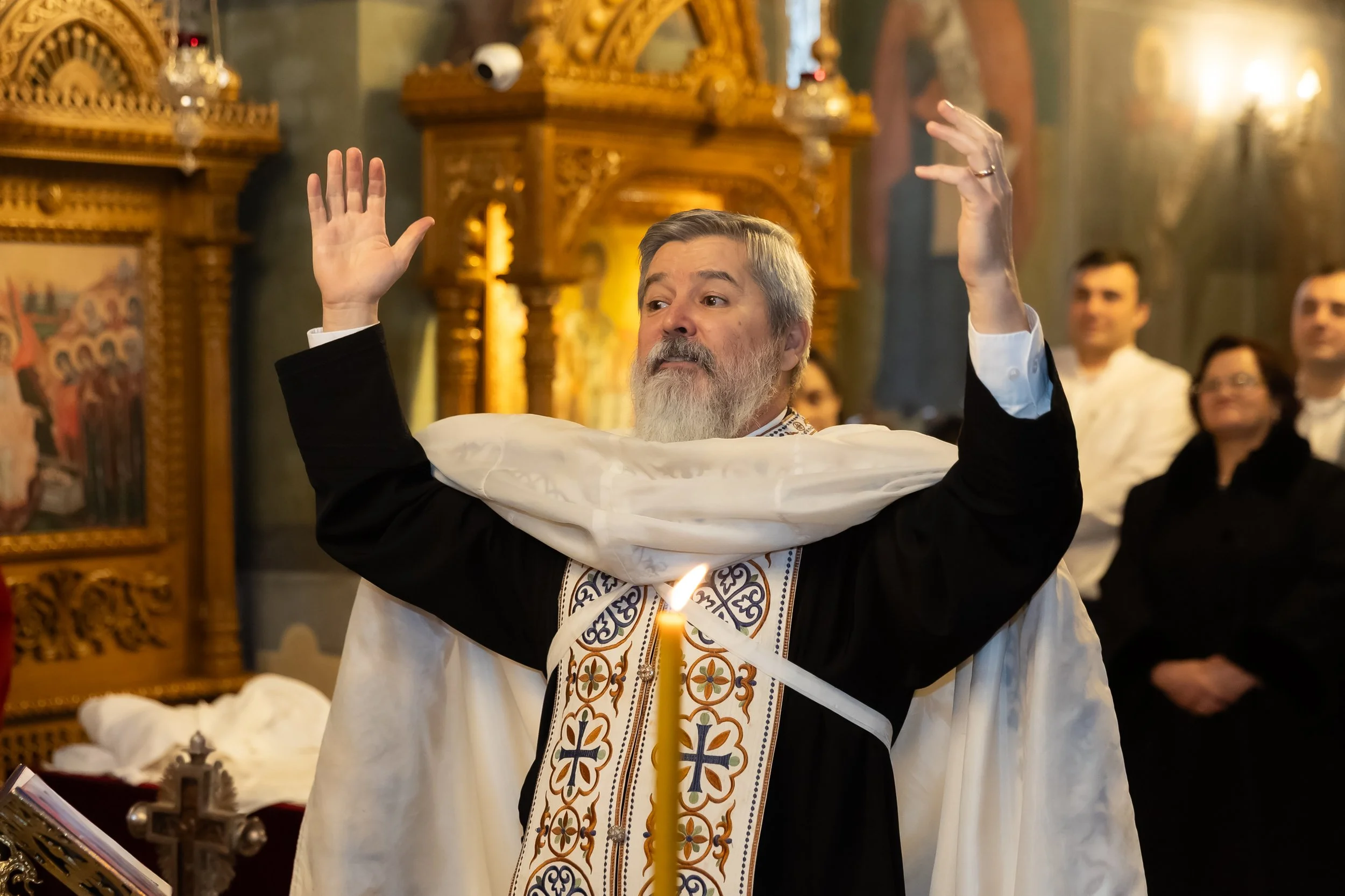 A priest with gray hair and a beard performing a religious ceremony inside a church, raising his hands. There are people watching him in the background. The church has ornate wooden decorations and religious icons.