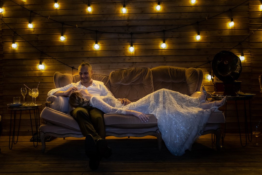 A couple lying on a beige sofa under warm string lights in a cozy, wooden room, with drinks on nearby tables.