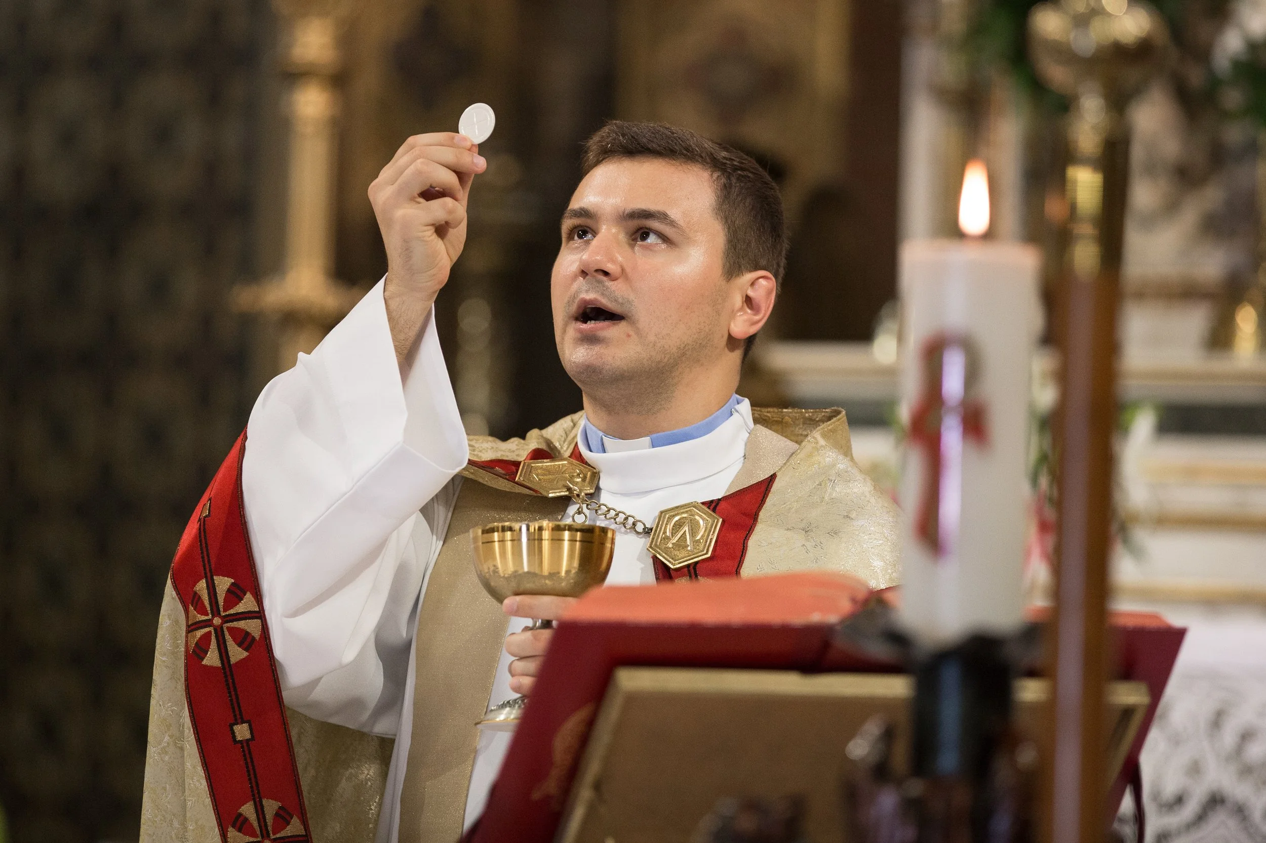 A man dressed as a priest or religious figure holding a chalice during a religious ceremony inside a church, with candles visible in the background.