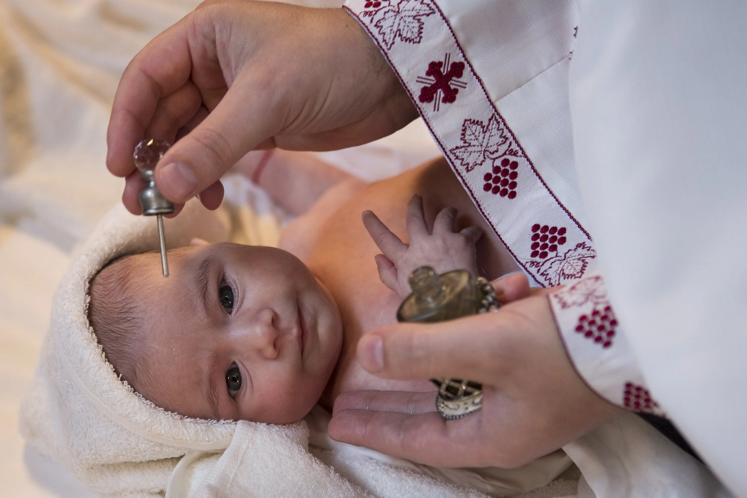 A baby with blue eyes lying wrapped in a towel while a person in a white embroidered garment gently holds the baby's hand during a religious or ceremonial act.