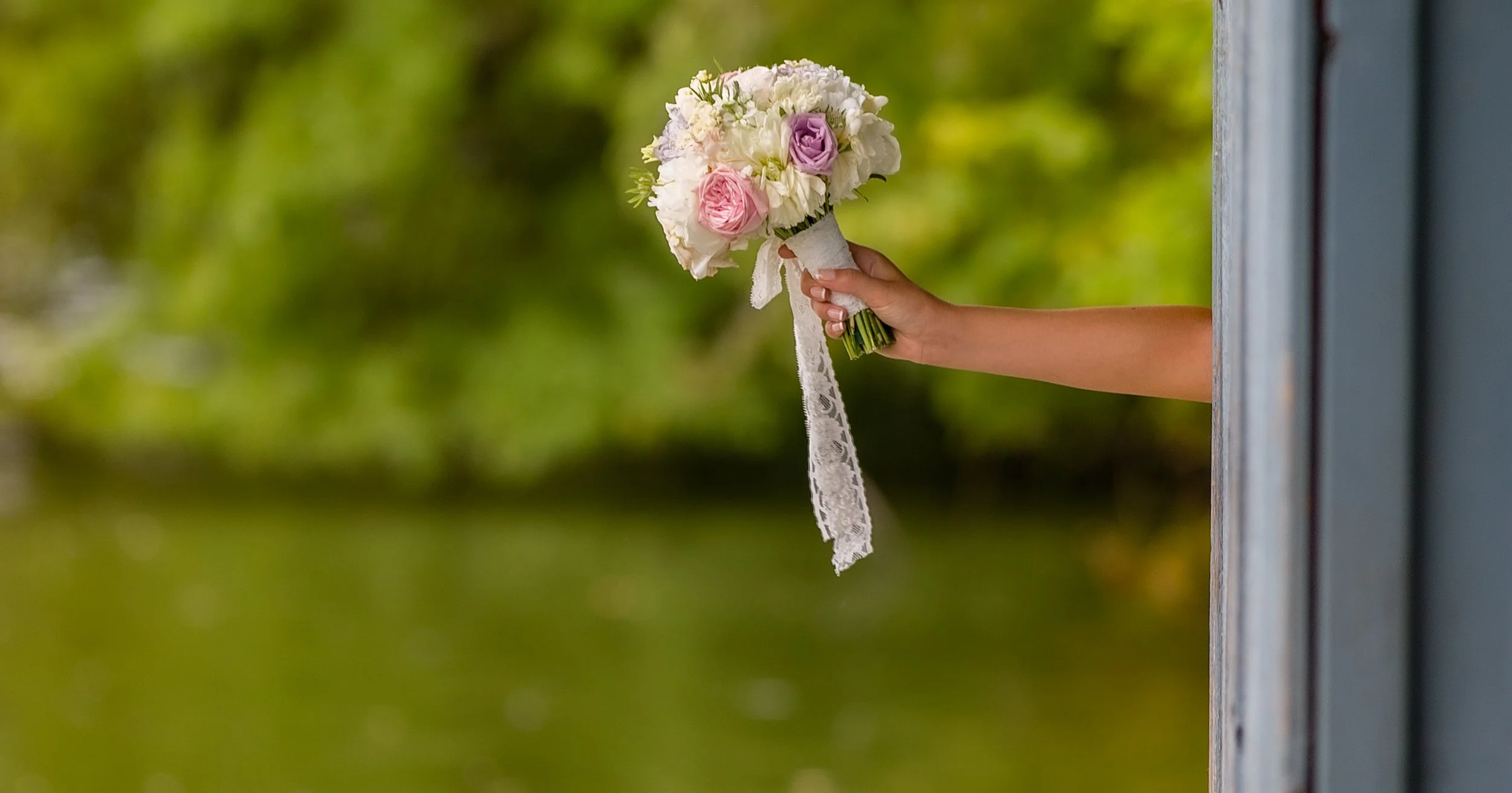 A person is holding a bouquet of flowers outside a window, extending it through the window opening. The bouquet includes pink and purple roses with white flowers and a lace ribbon. The background is blurred greenery.