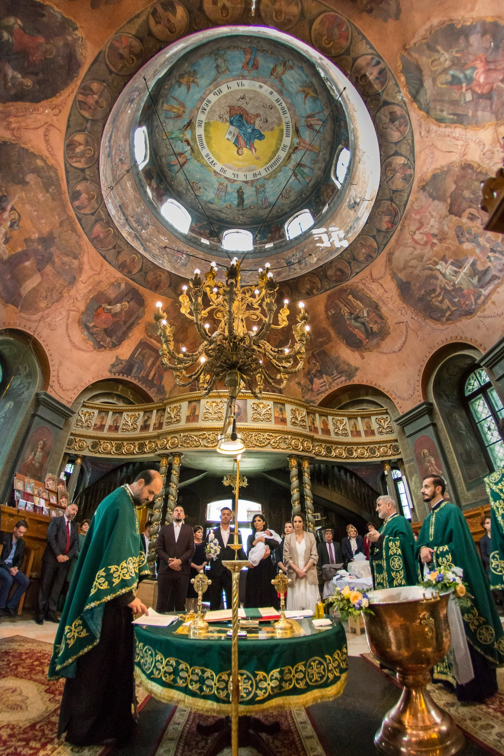Inside of a church with a decorated ceiling and a large chandelier. A group of people, including clergy in green vestments, are gathered around a altar decorated with a green cloth and religious items, participating in a religious ceremony. The ceili