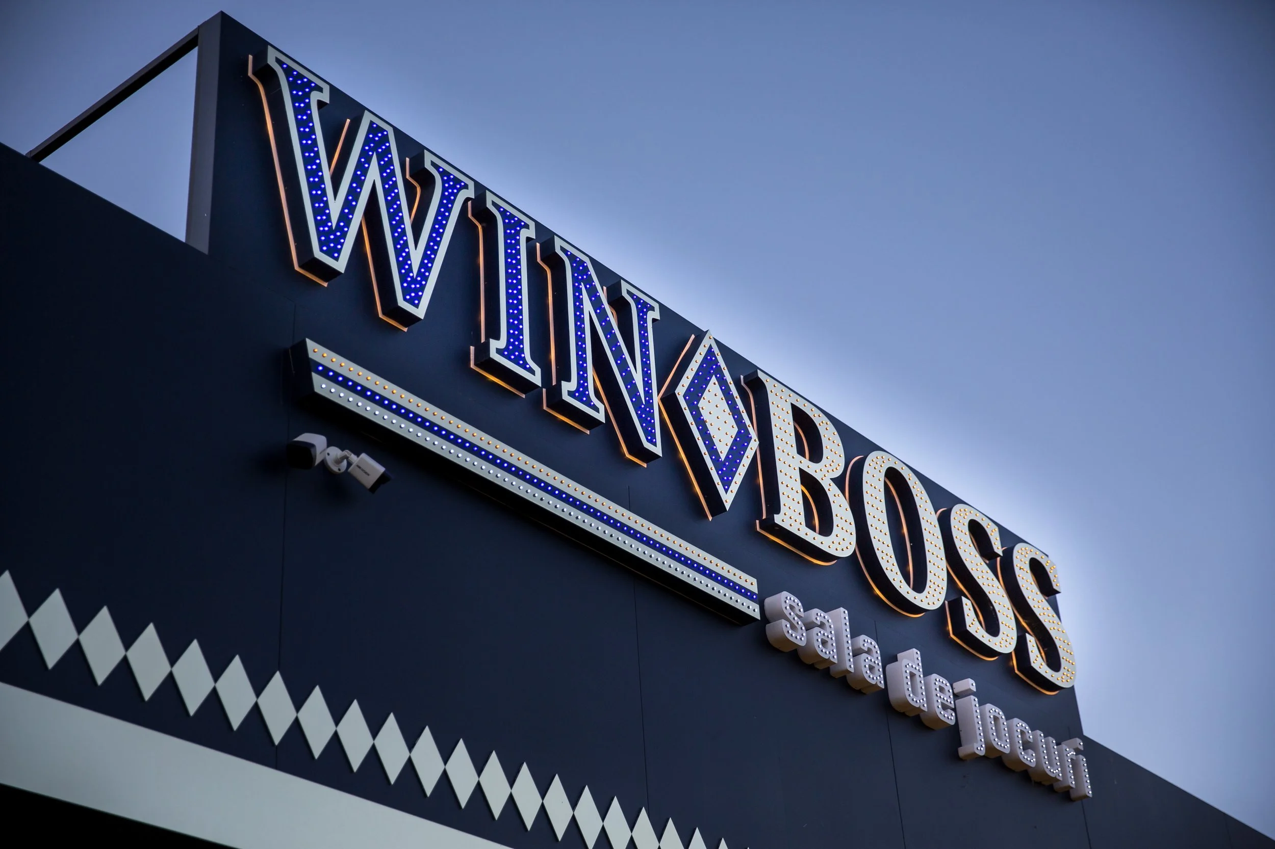 Close-up of a vintage-style illuminated sign with the word 'WINGS' in large letters, attached to the side of a building with a dark blue background, taken during dusk.