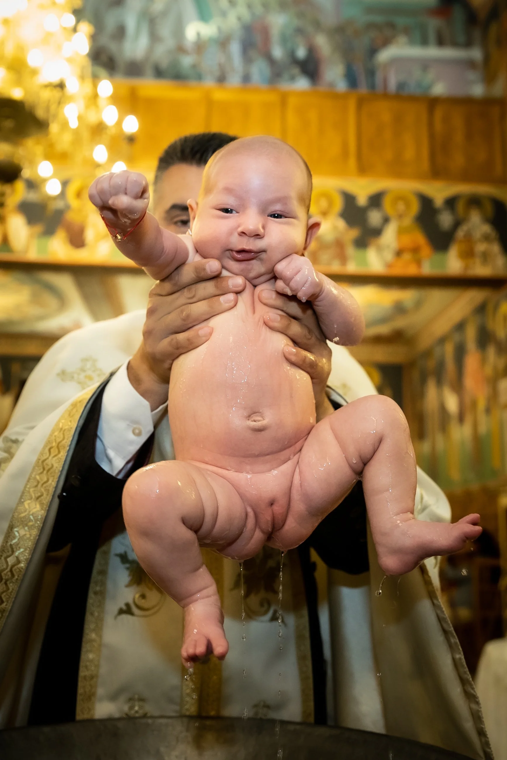 A person holding a baby during a religious baptism ceremony inside a church, with a decorated background and warm lighting.