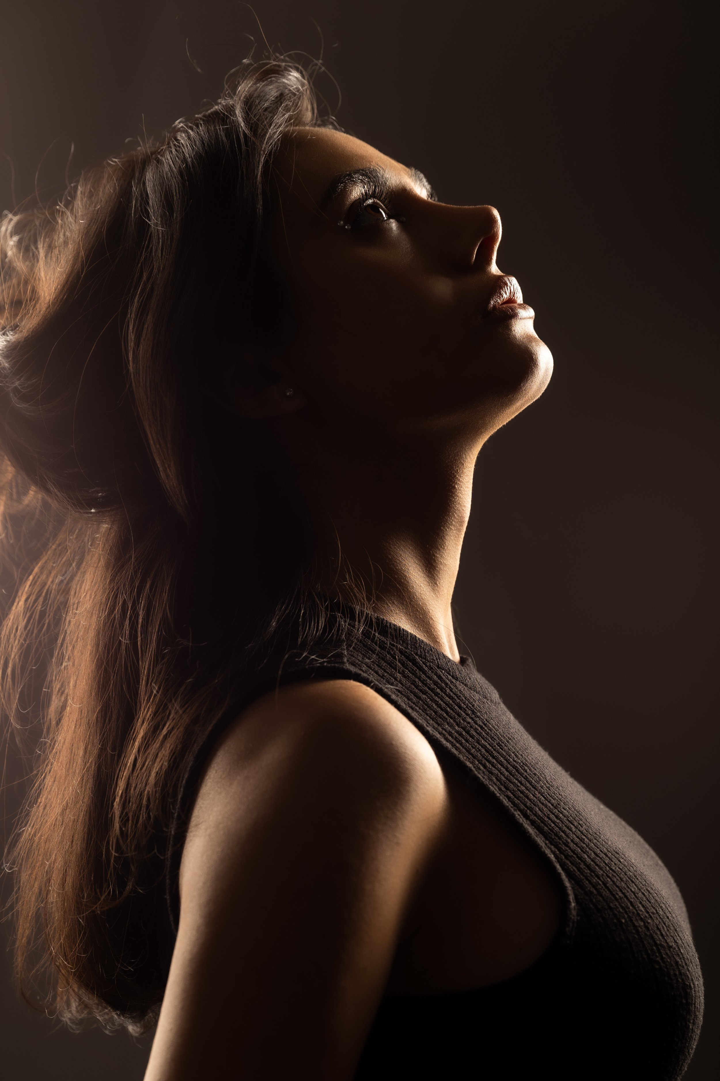 A woman with long hair gazing upward in profile, illuminated with soft, warm lighting, wearing a black sleeveless top.