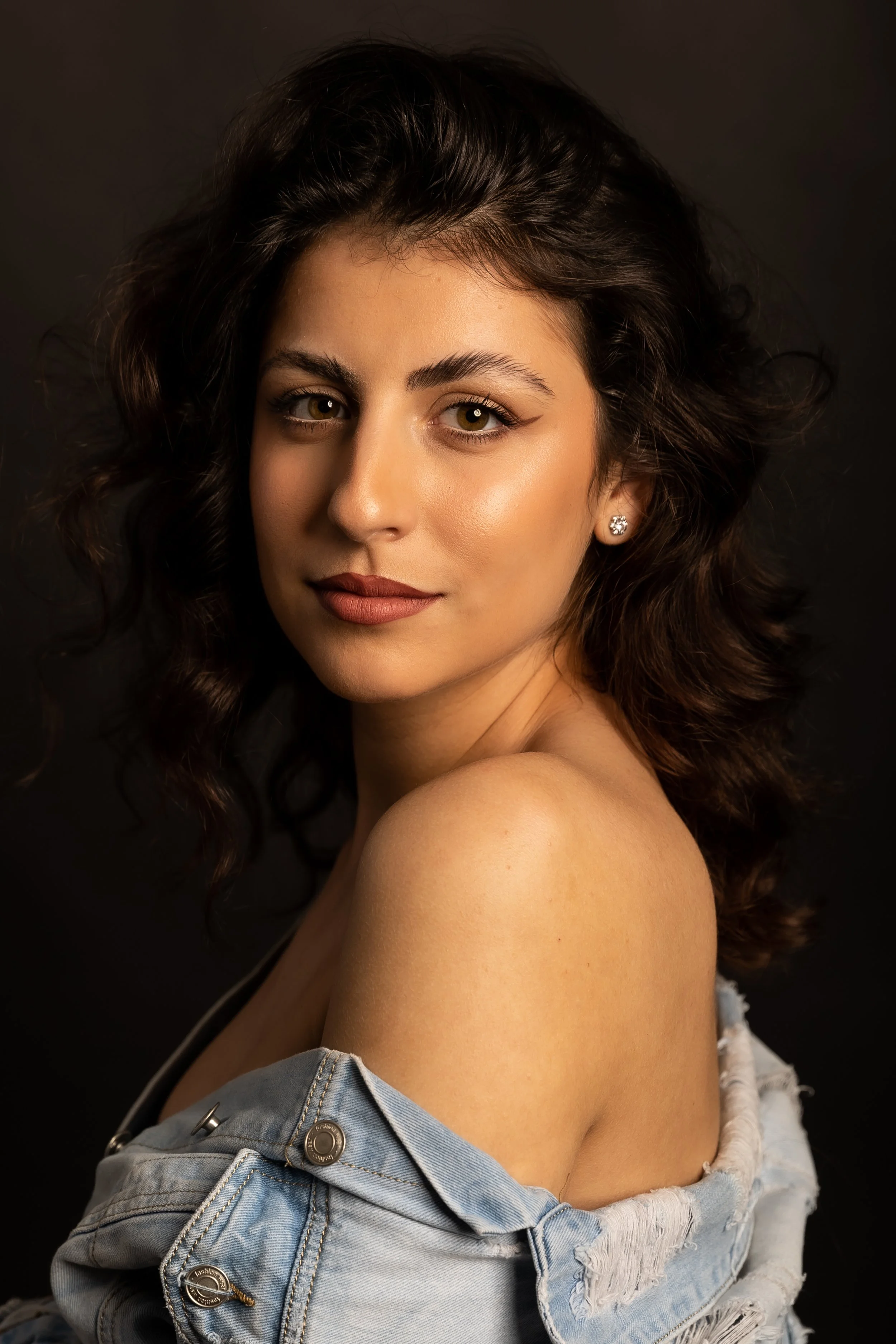Close-up of a young woman with wavy brown hair, hazel eyes, and light makeup, wearing a distressed denim jacket off one shoulder, against a dark background.