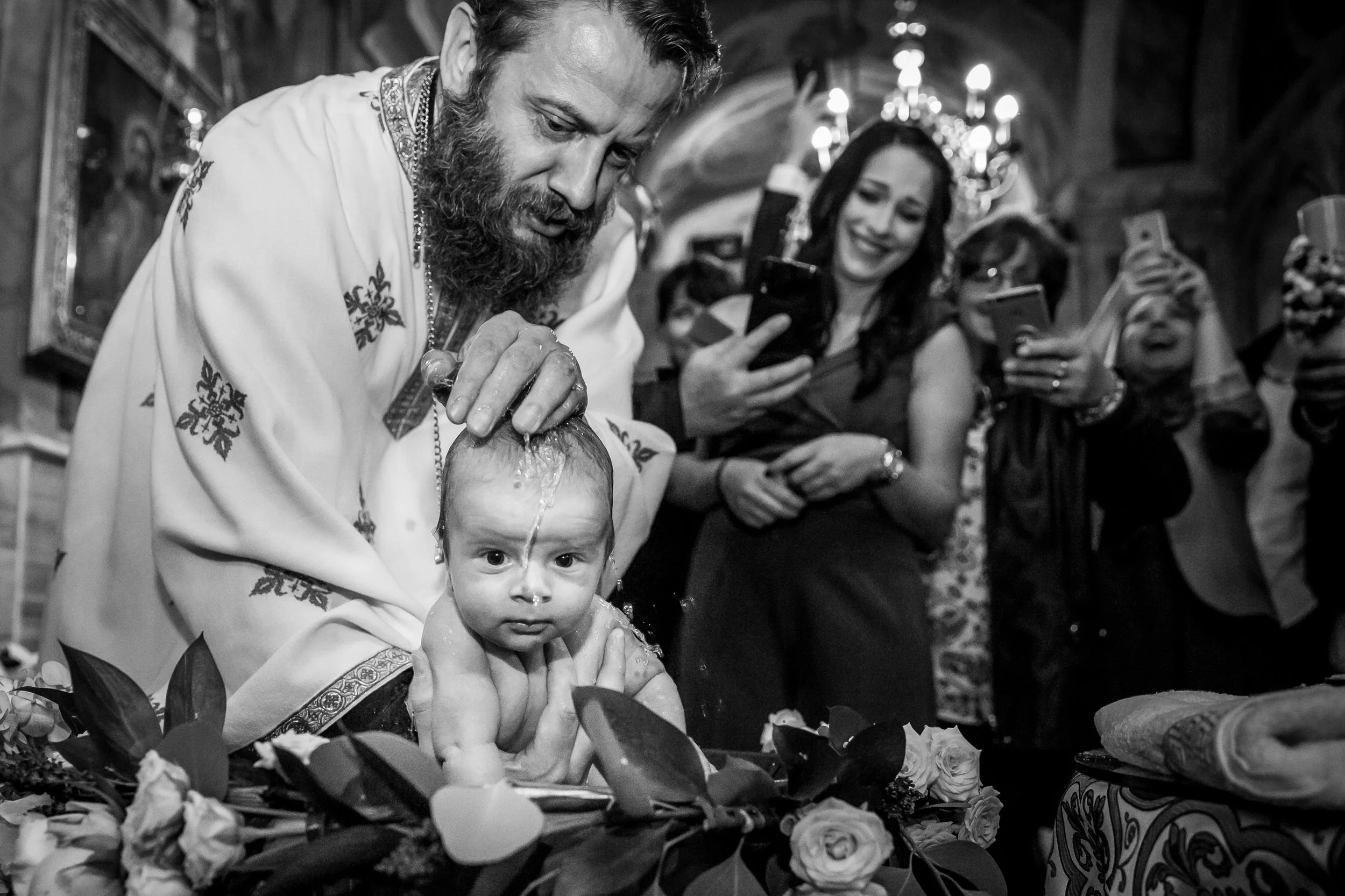 A religious ceremony where a priest is performing a baptism or blessing on a baby, surrounded by a group of people taking photos and observing.
