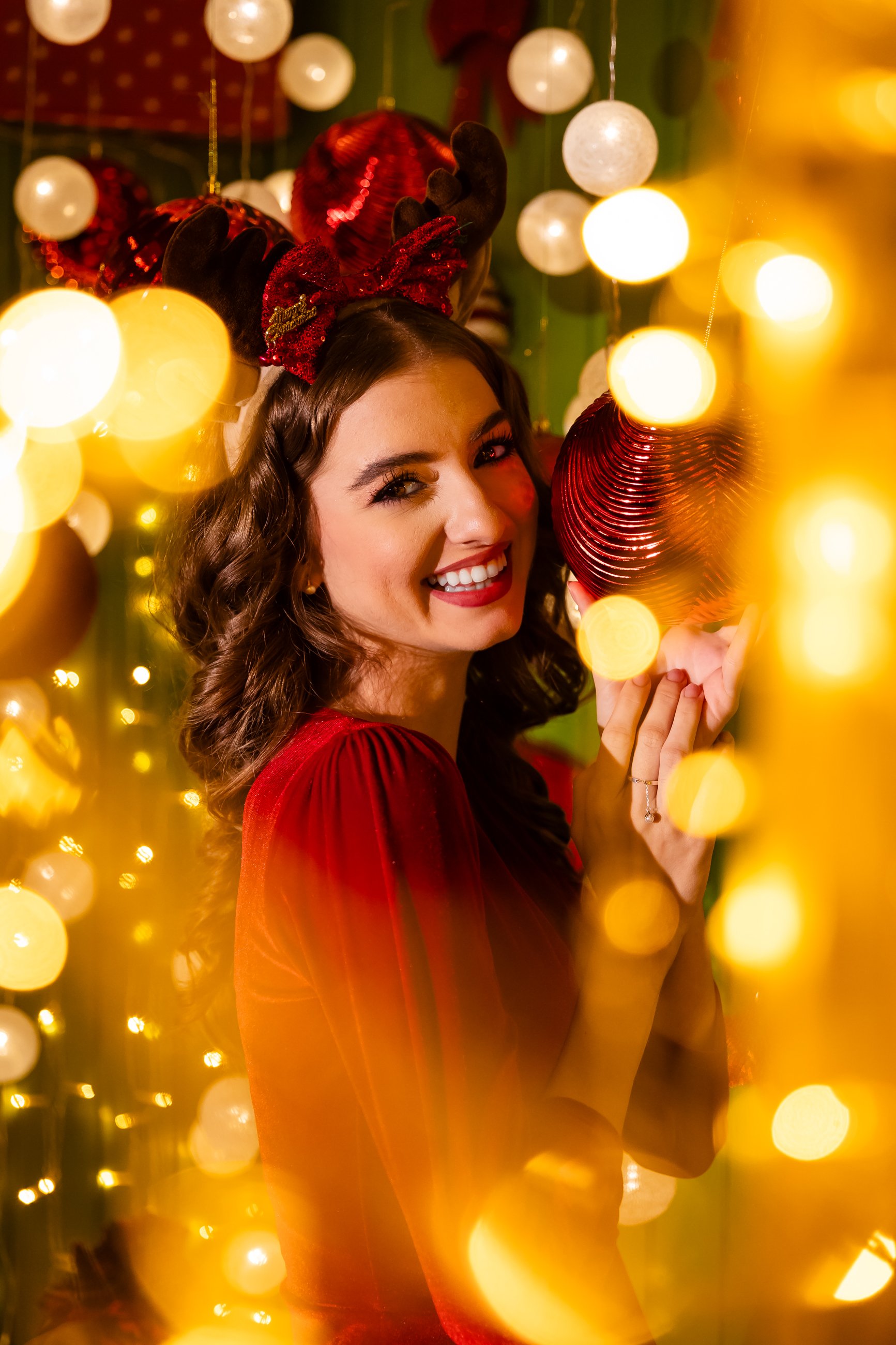 A woman with curly hair wearing reindeer antlers headband and a red dress, smiling near Christmas decorations with glowing lights and ornaments.