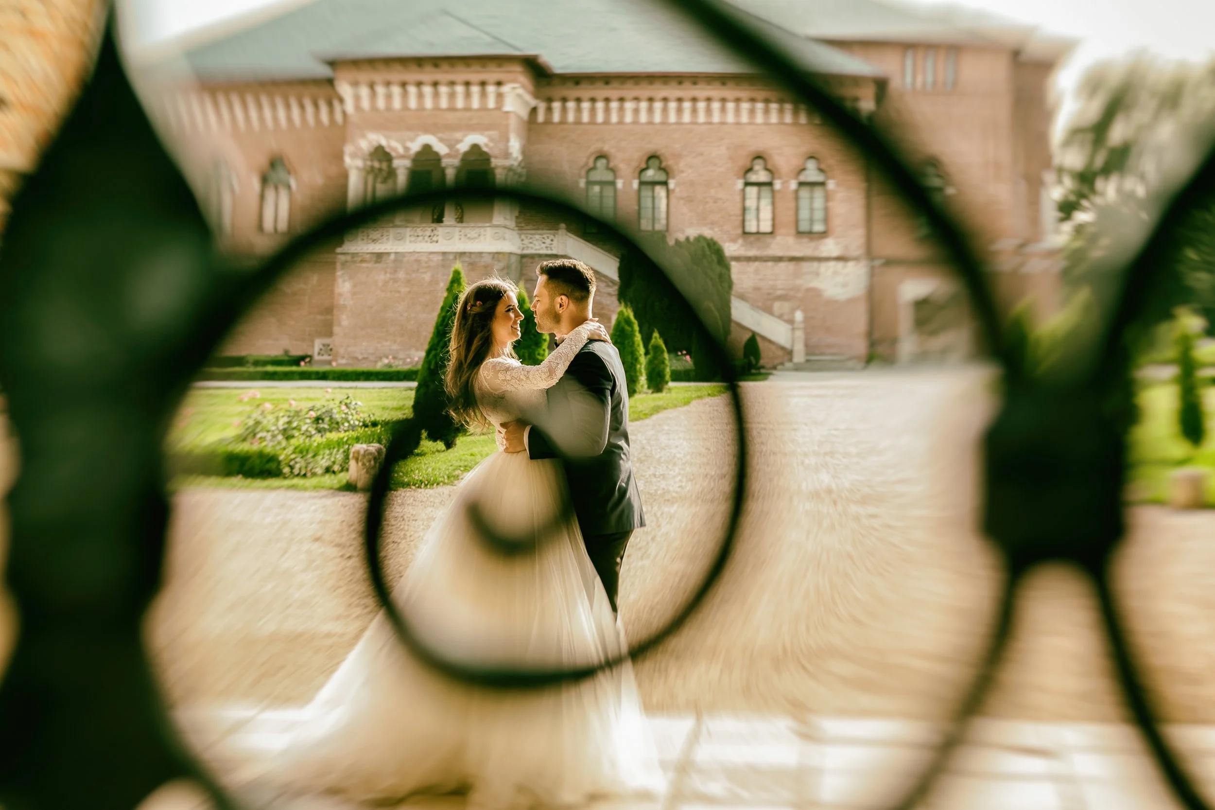 A couple dressed in wedding attire, embracing and smiling at each other, seen through a decorative iron fence with a historic brick building in the background.