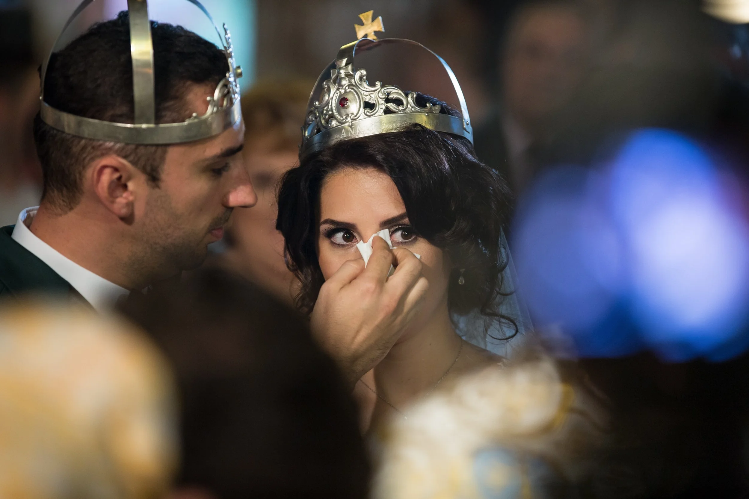 A woman with dark curly hair receives a tissue wipe from a man wearing a silver crown, likely during a ceremonial event.