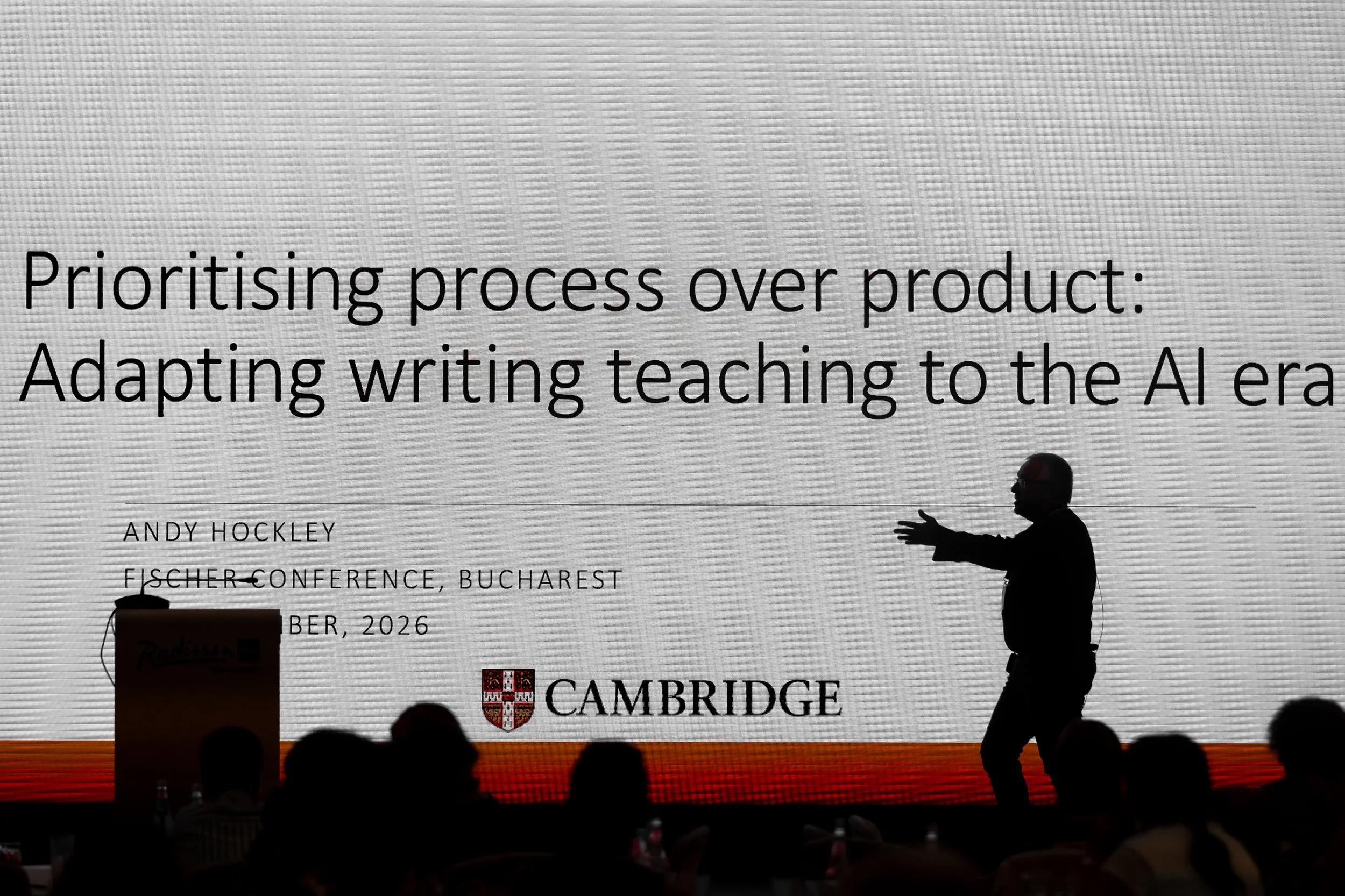 Group of people sitting in an auditorium facing a large presentation screen. The screen displays a slide titled 'Prioritising process over product: Adapting writing teaching to the Aler', with information about Andy Hockley, Fischer Conference, Bucha