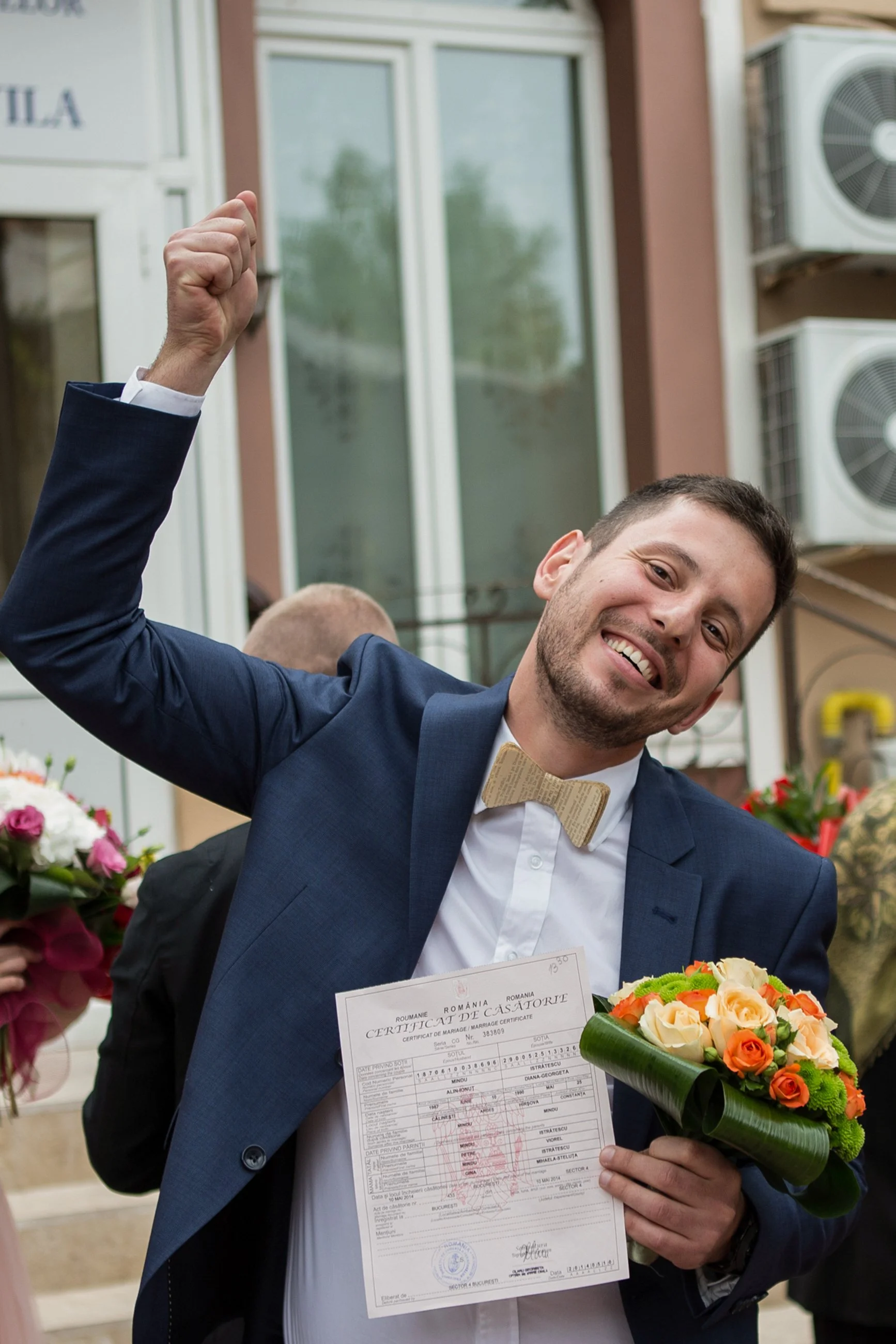 A man wearing a dark blue suit and a beige bow tie, smiling and holding a marriage certificate and a bouquet of flowers, with a background of a building and air conditioning units.