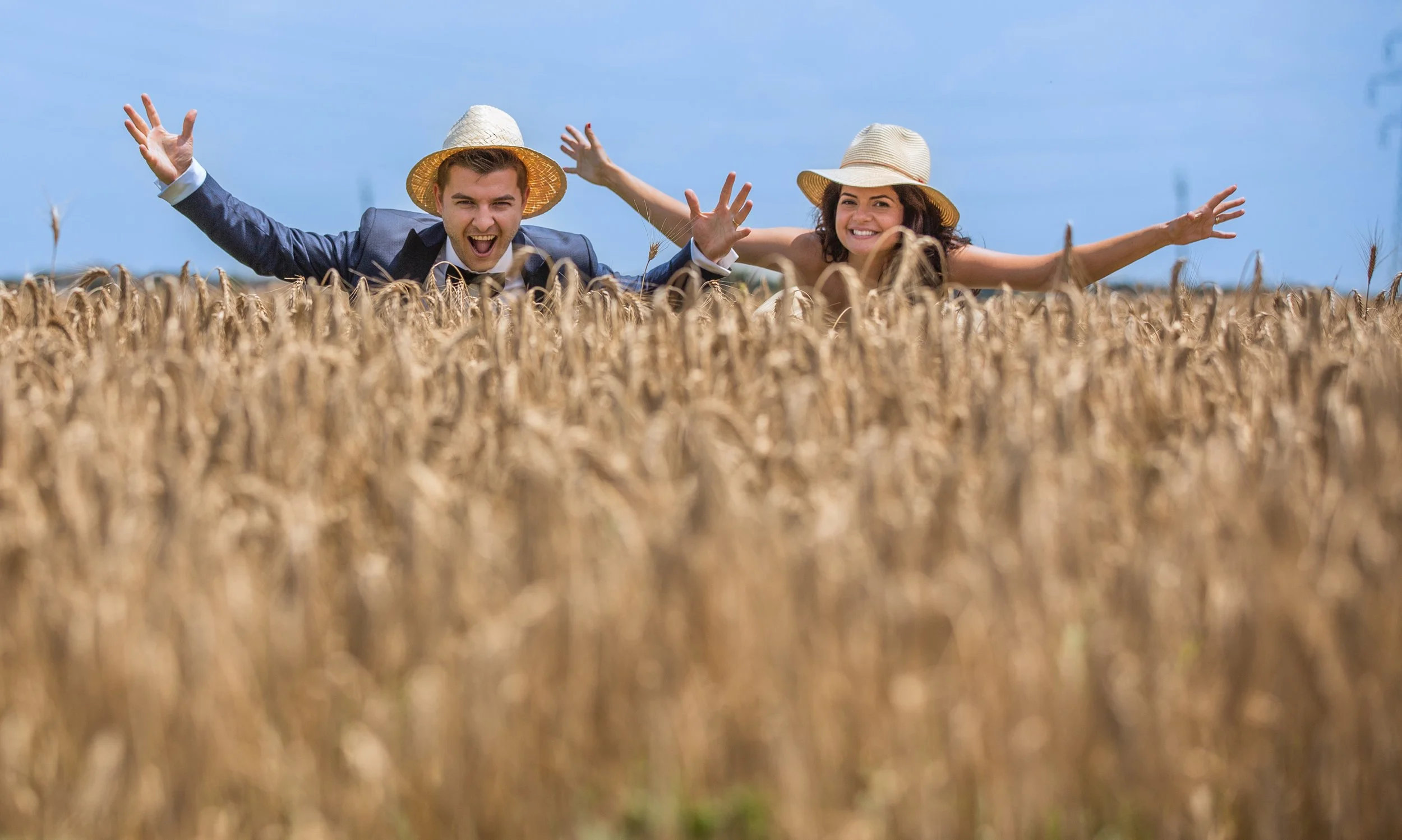 A man and woman wearing straw hats and formal attire, smiling and stretching their arms out in front of a wheat field during daytime.