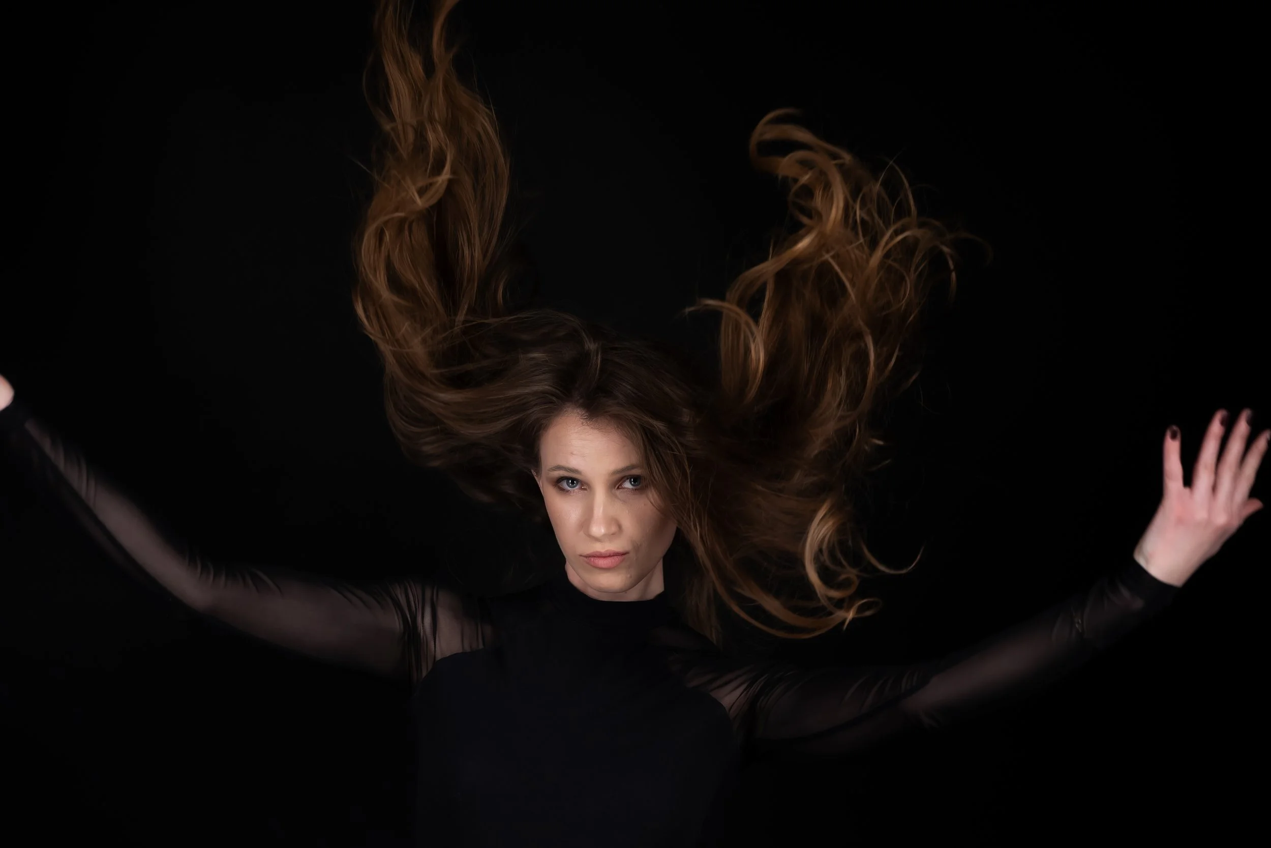A woman with long, wavy red hair spread out above her head, wearing black sheer clothing, looking directly at the camera against a black background.