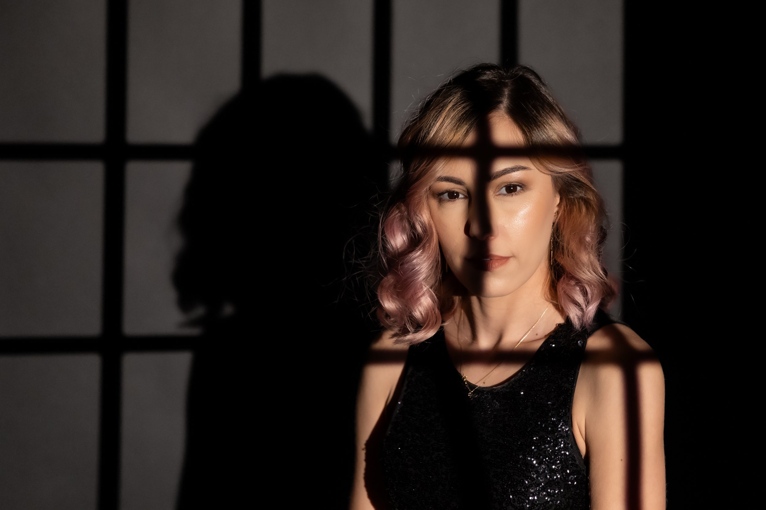 A young woman with wavy pink and blonde hair, wearing a black sequin dress, is looking directly at the camera behind bars casting shadows on her face.