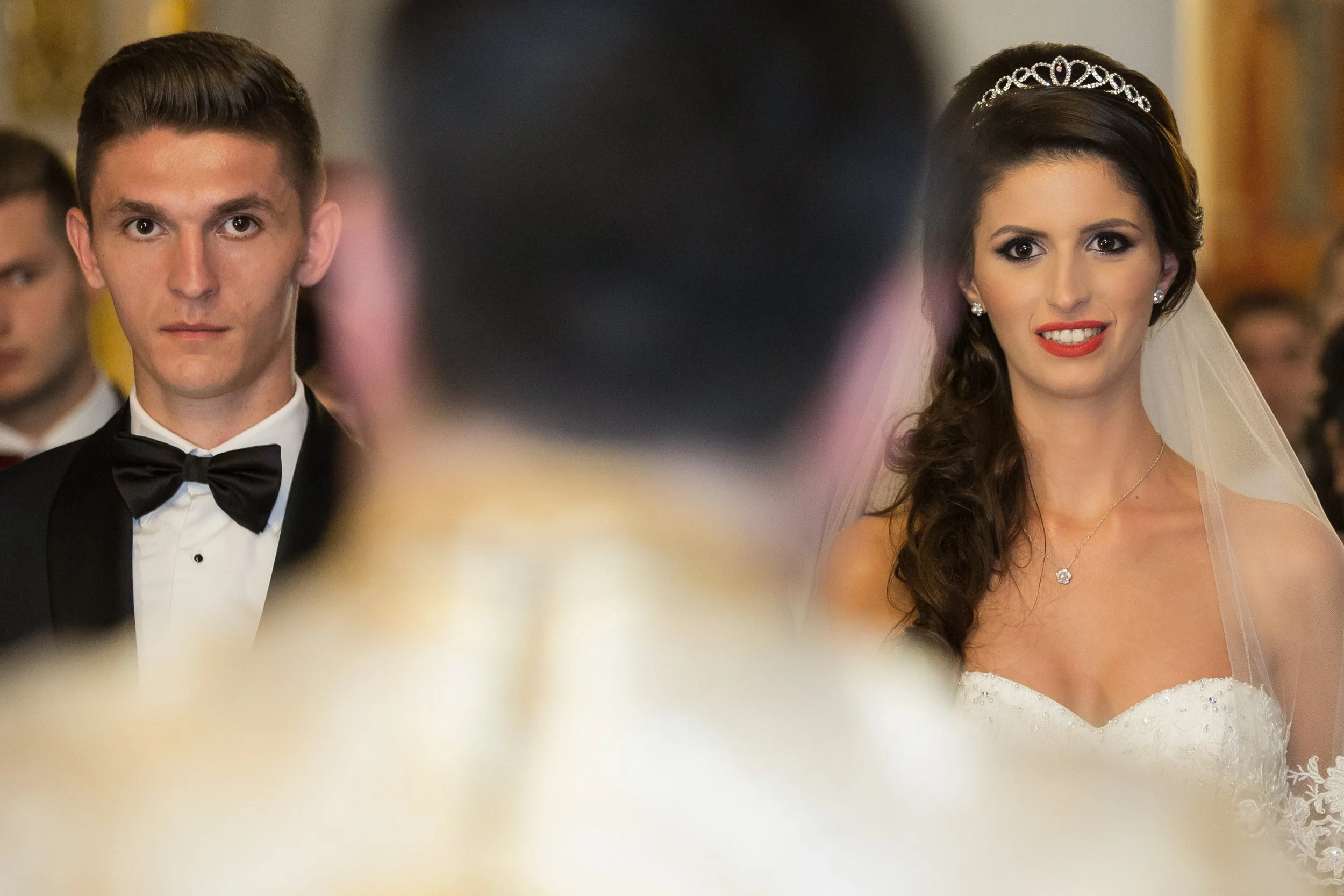 A bride in a white wedding dress with lace details and a tiara, and a groom in a black tuxedo with a bow tie, during a wedding ceremony in a church or reception hall.