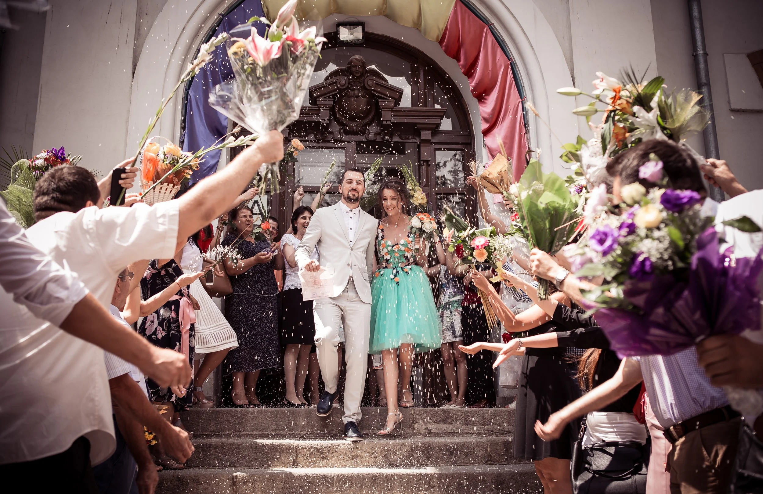 A newlywed couple, dressed in wedding attire, walking down the steps of a building while being celebrated by friends and family who are throwing flower petals and confetti. The scene is festive with colorful flowers and decorations.