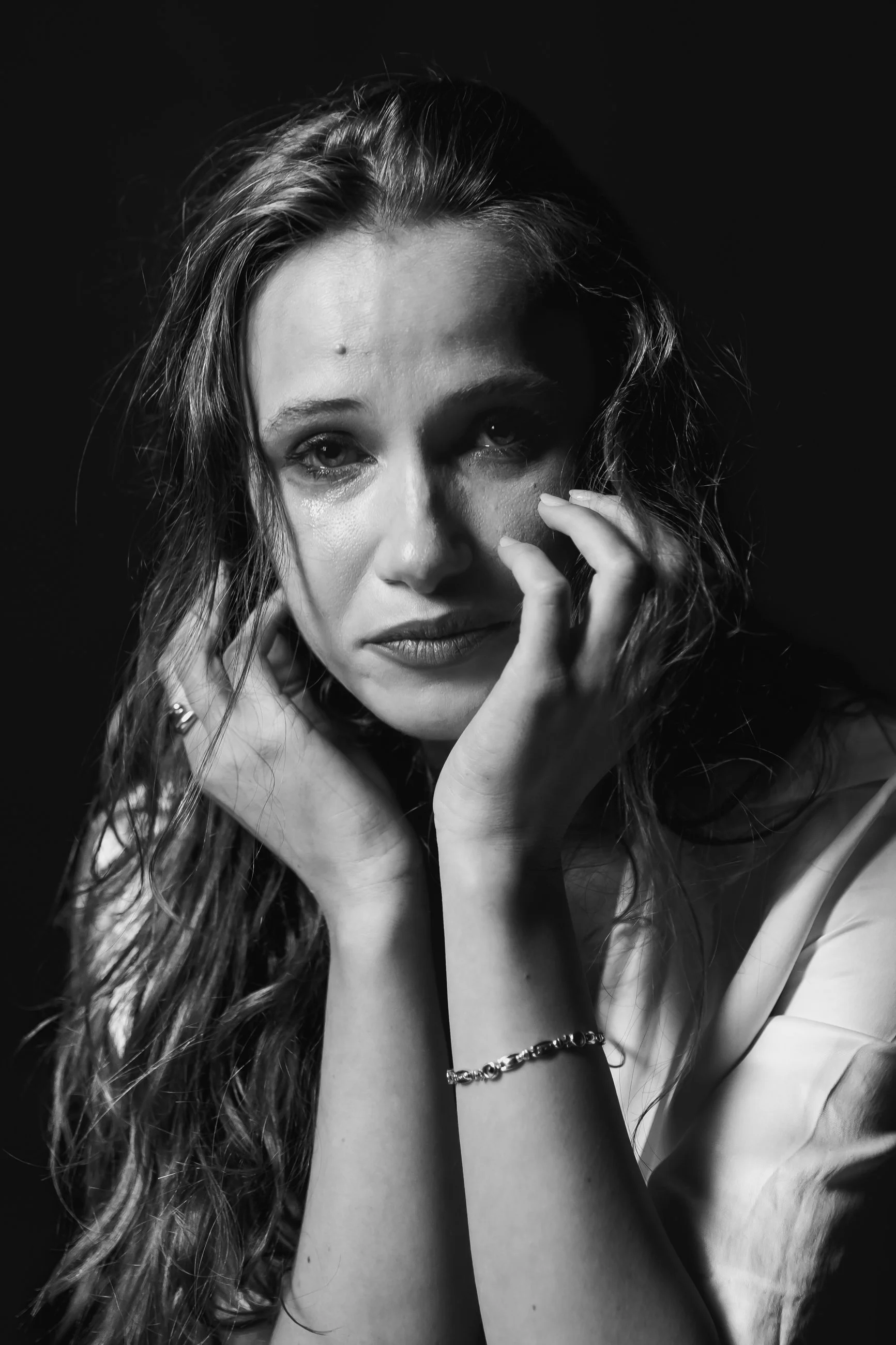 Black and white portrait of a woman with long wavy hair, touching her face and neck, with one eye slightly open and tears visible, against a dark background.