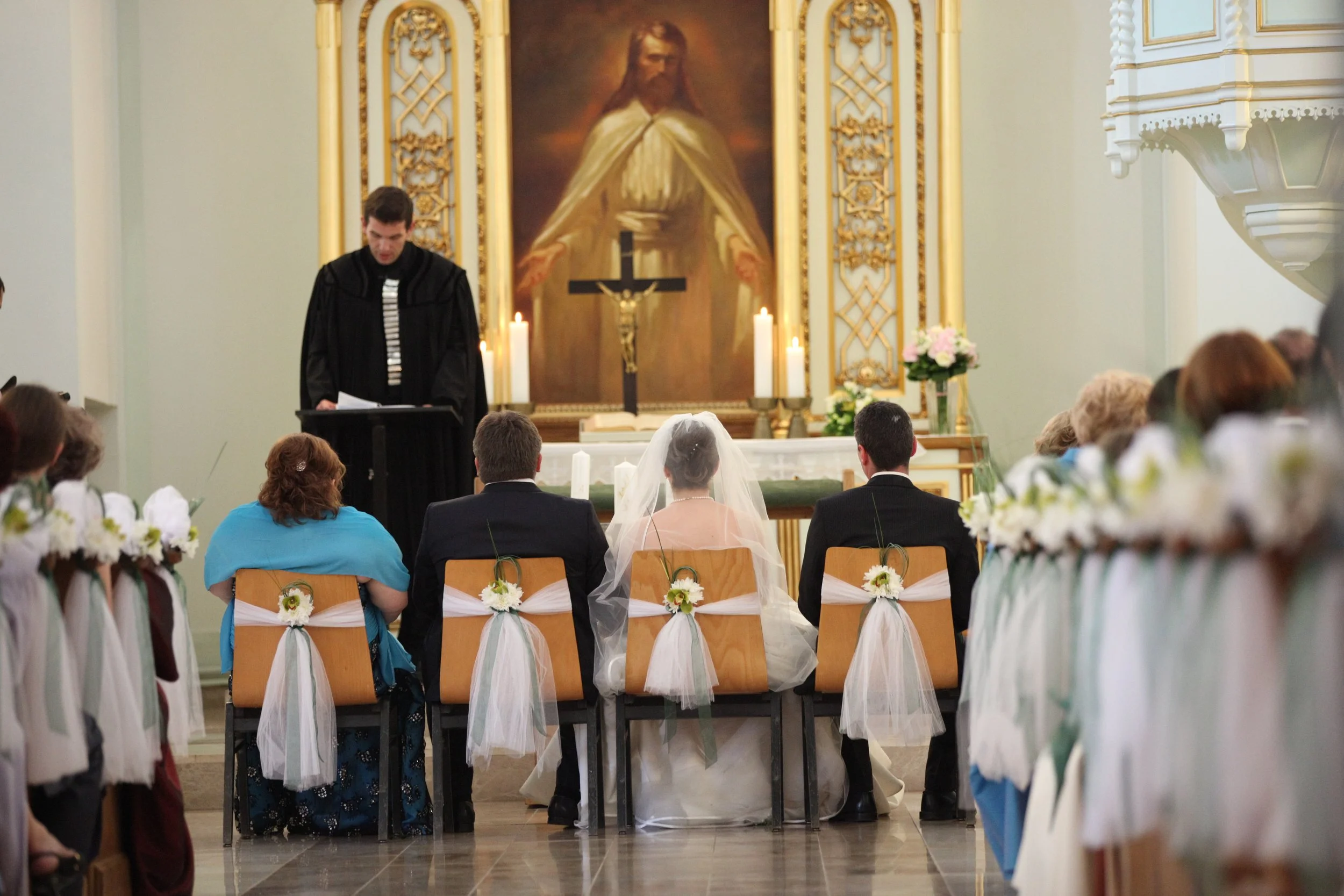 A couple sitting in front of an altar during a wedding ceremony in a church, with a priest or officiant standing at a lectern. The church has a large portrait of Jesus Christ on the wall behind the altar, lit candles, and floral decorations on the pe
