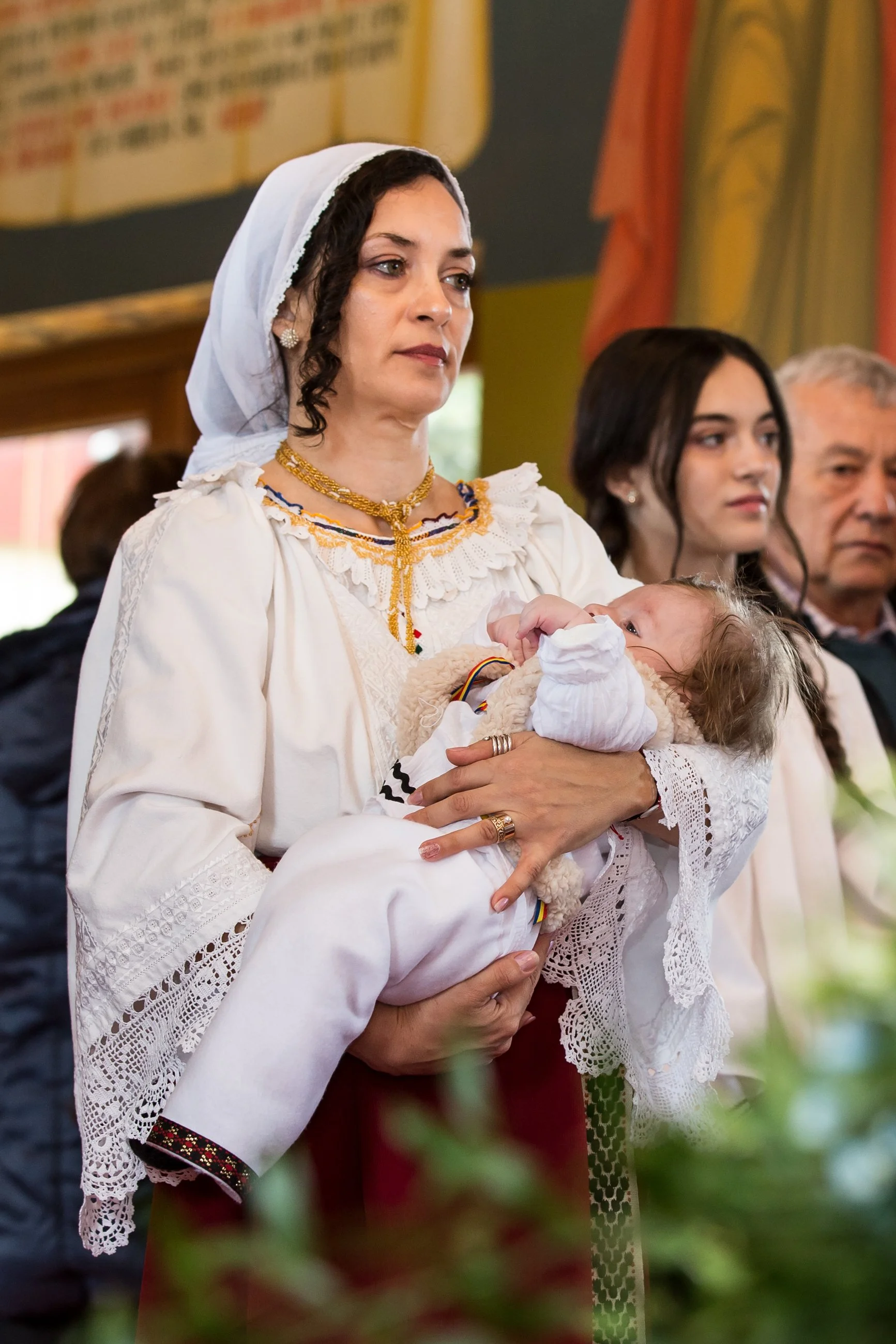 A woman in traditional dress holding a baby during a ceremony, with other people standing behind her.