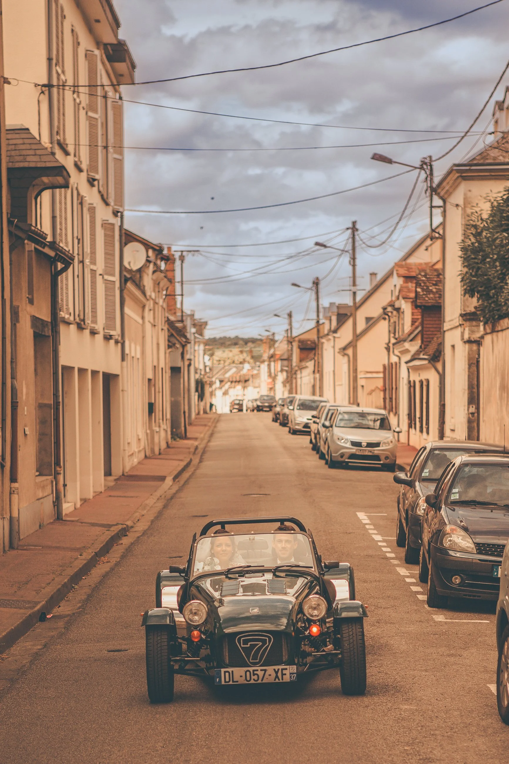 A vintage black sports car with two passengers driving down a narrow city street lined with parked cars and European-style buildings under a cloudy sky.