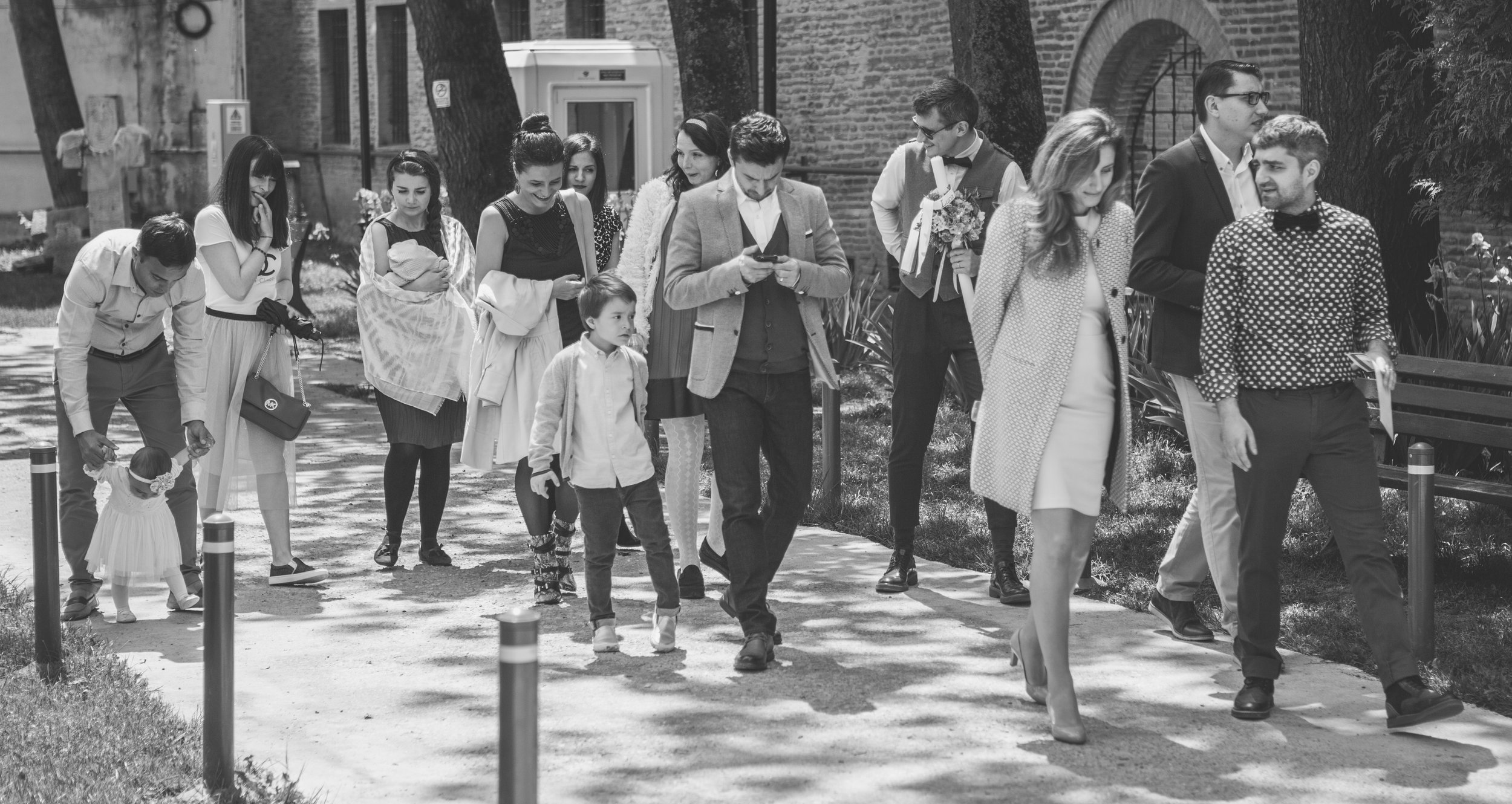 A group of people, including children and adults, walking along a sidewalk outdoors with brick buildings and trees in the background.