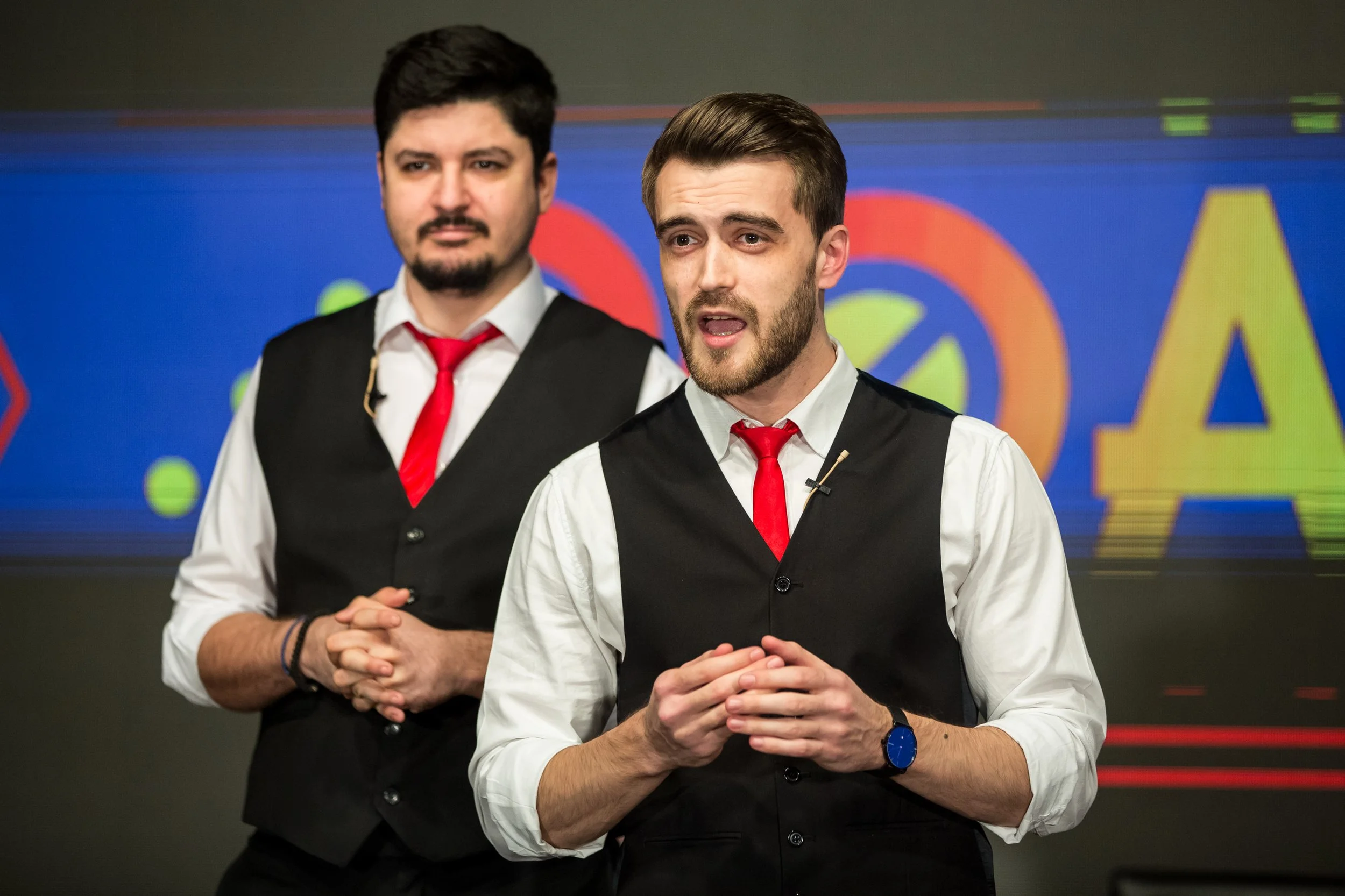 Two men in dress shirts and vests with red ties standing in front of a colorful background, possibly at a presentation or event.