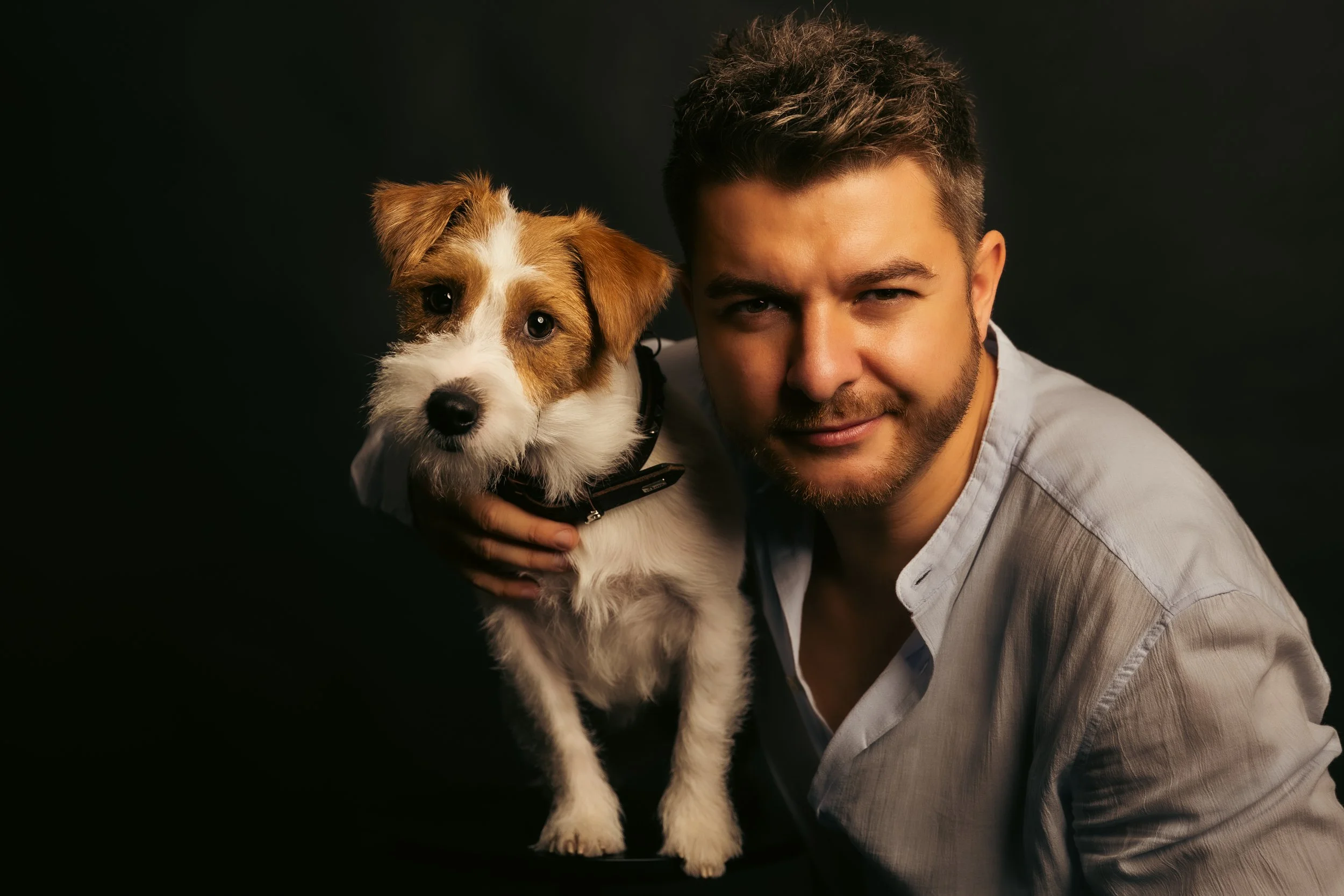 A man with dark hair and a beard holding a small brown and white puppy with floppy ears against a black background.