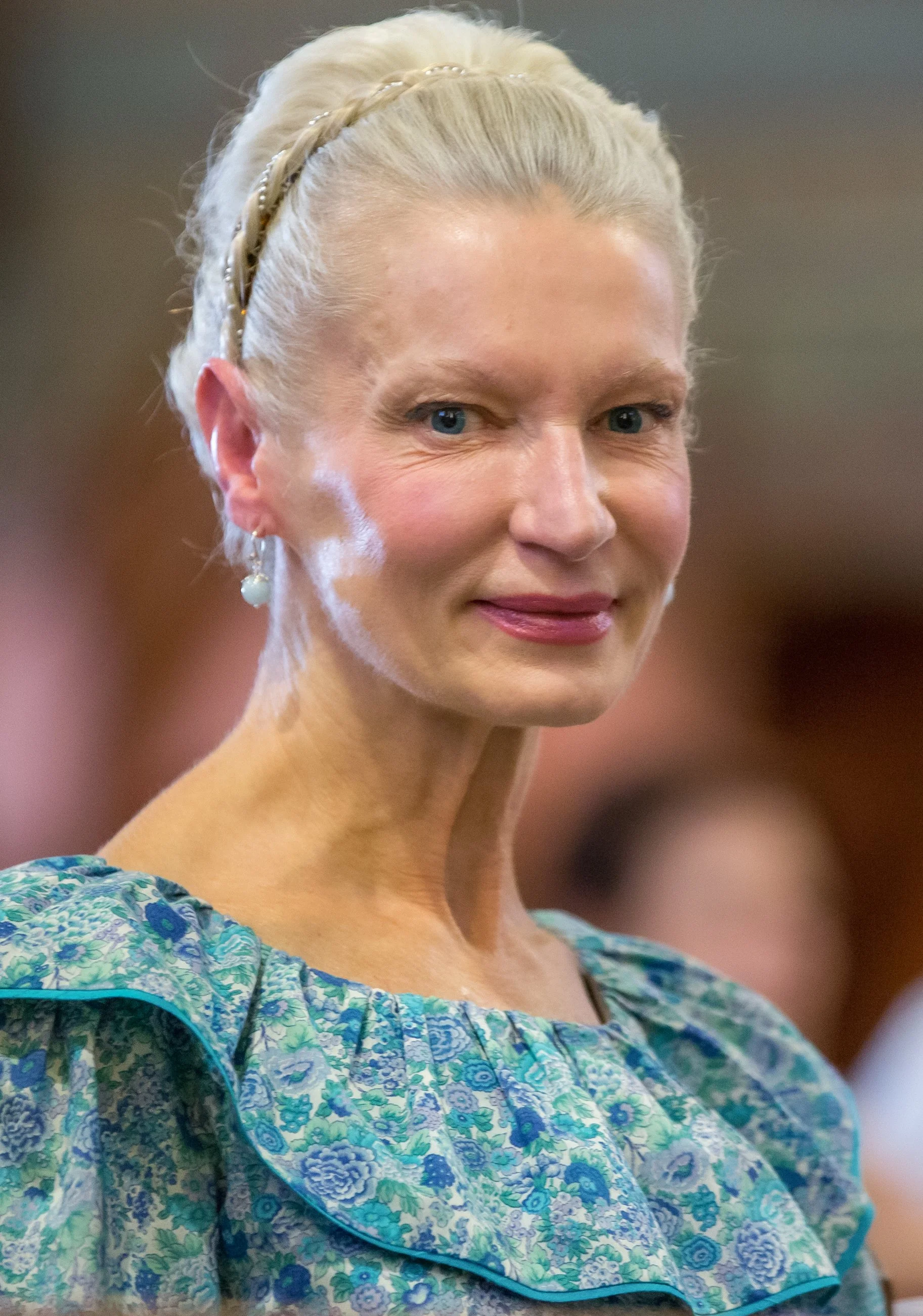 Close-up of a woman with platinum blonde hair styled in an updo with a braided headband, wearing pearl earrings, and a floral dress with ruffled sleeves.