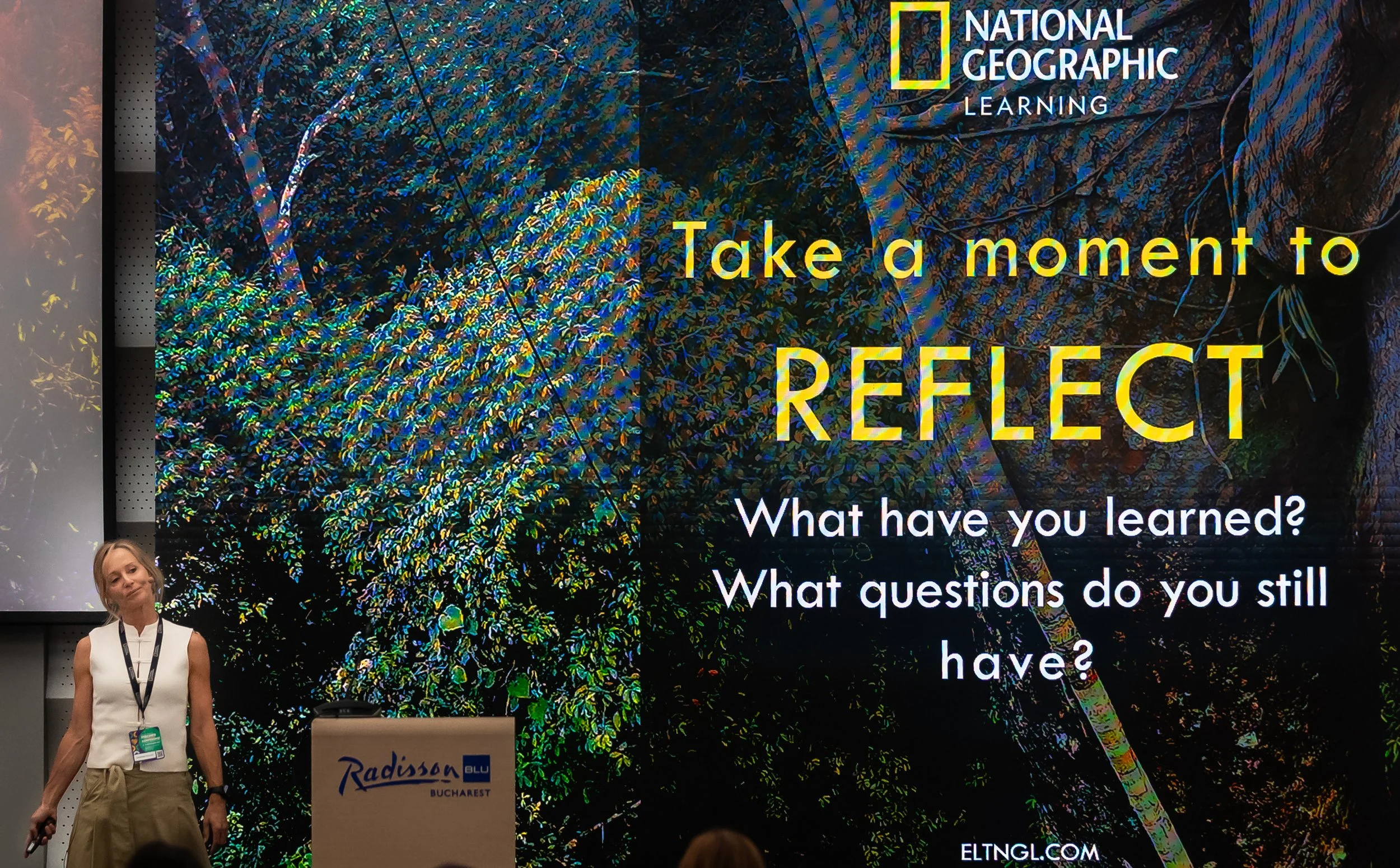 A woman standing in front of a large colorful digital display promoting a National Geographic Learning event with the phrase 'Take a moment to REFLECT'. The woman is holding a leash and has blonde hair, wearing a sleeveless white top and khaki pants.