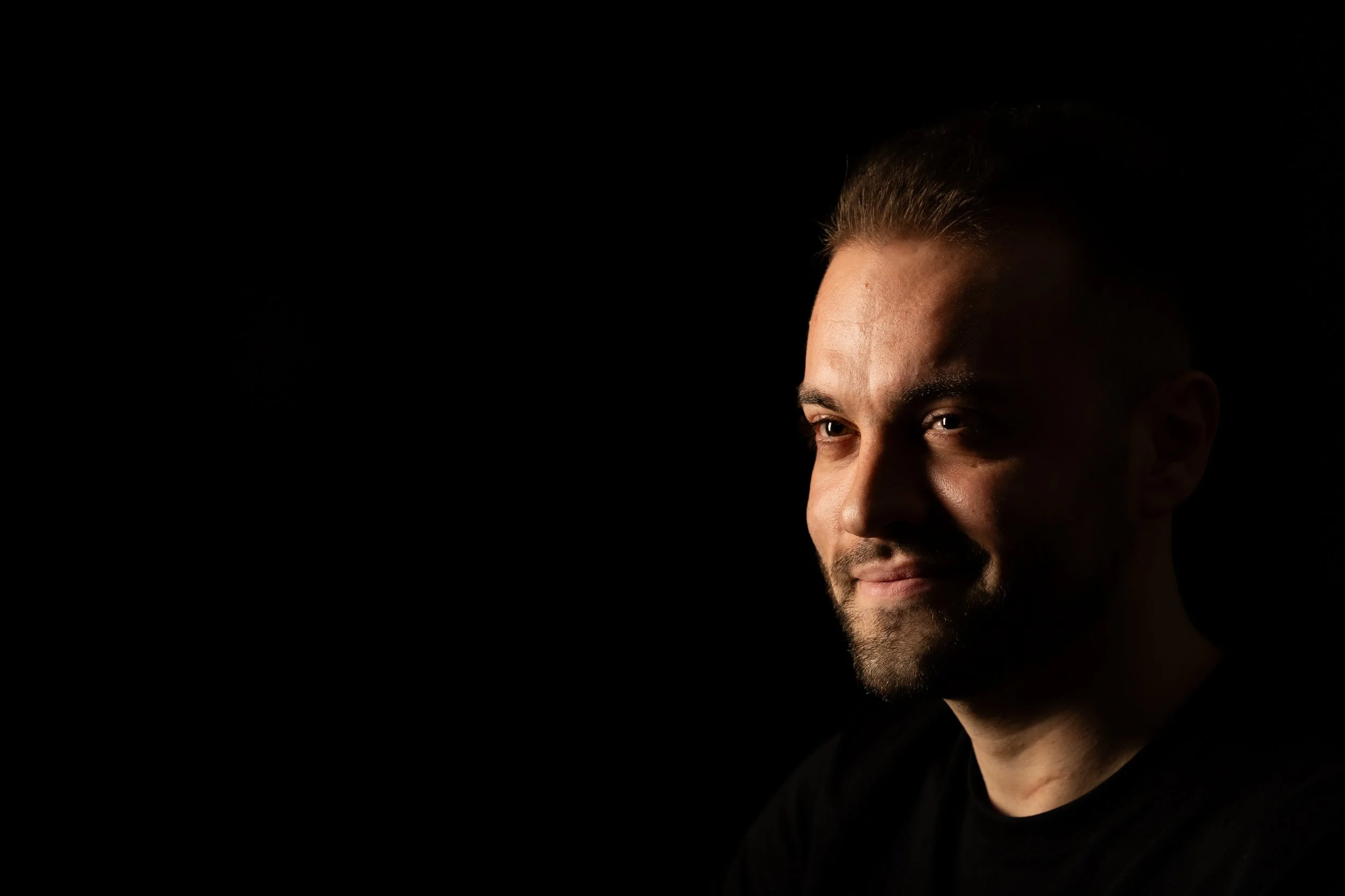 Close-up of a smiling man with short hair and a beard, looking to the side against a black background.
