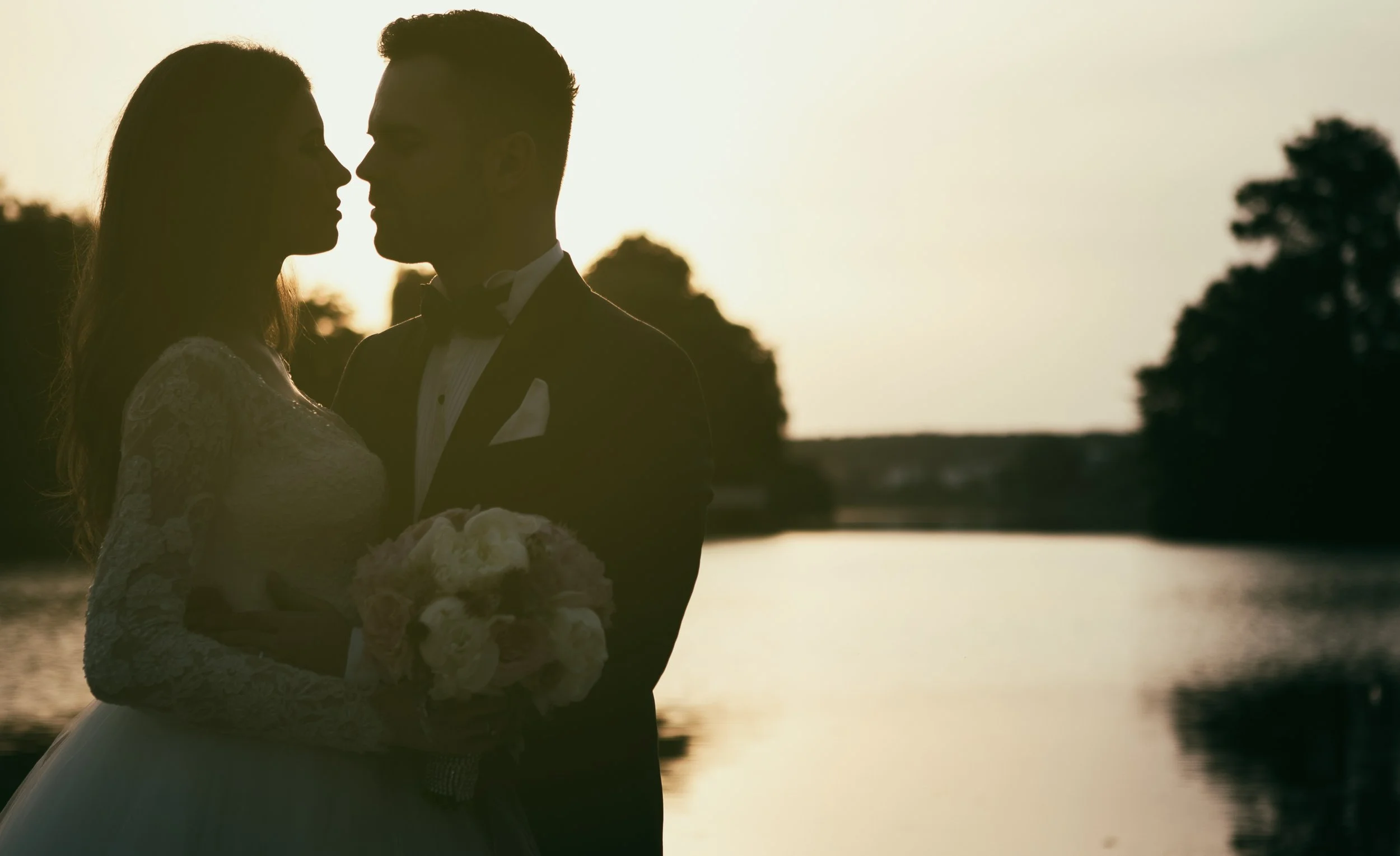 A silhouette of a bride and groom standing close together at sunset near a body of water, with trees in the background.