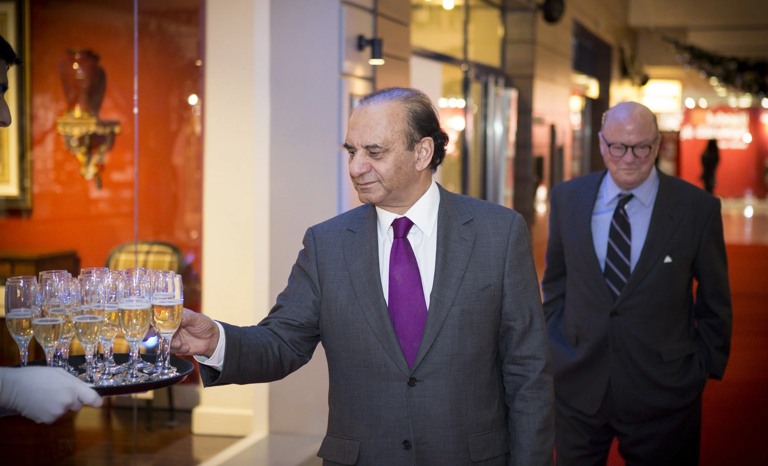 A man in a gray suit with purple tie holding a tray of champagne glasses at an event, with another man in a dark suit and glasses walking behind him in a decorated indoor setting.