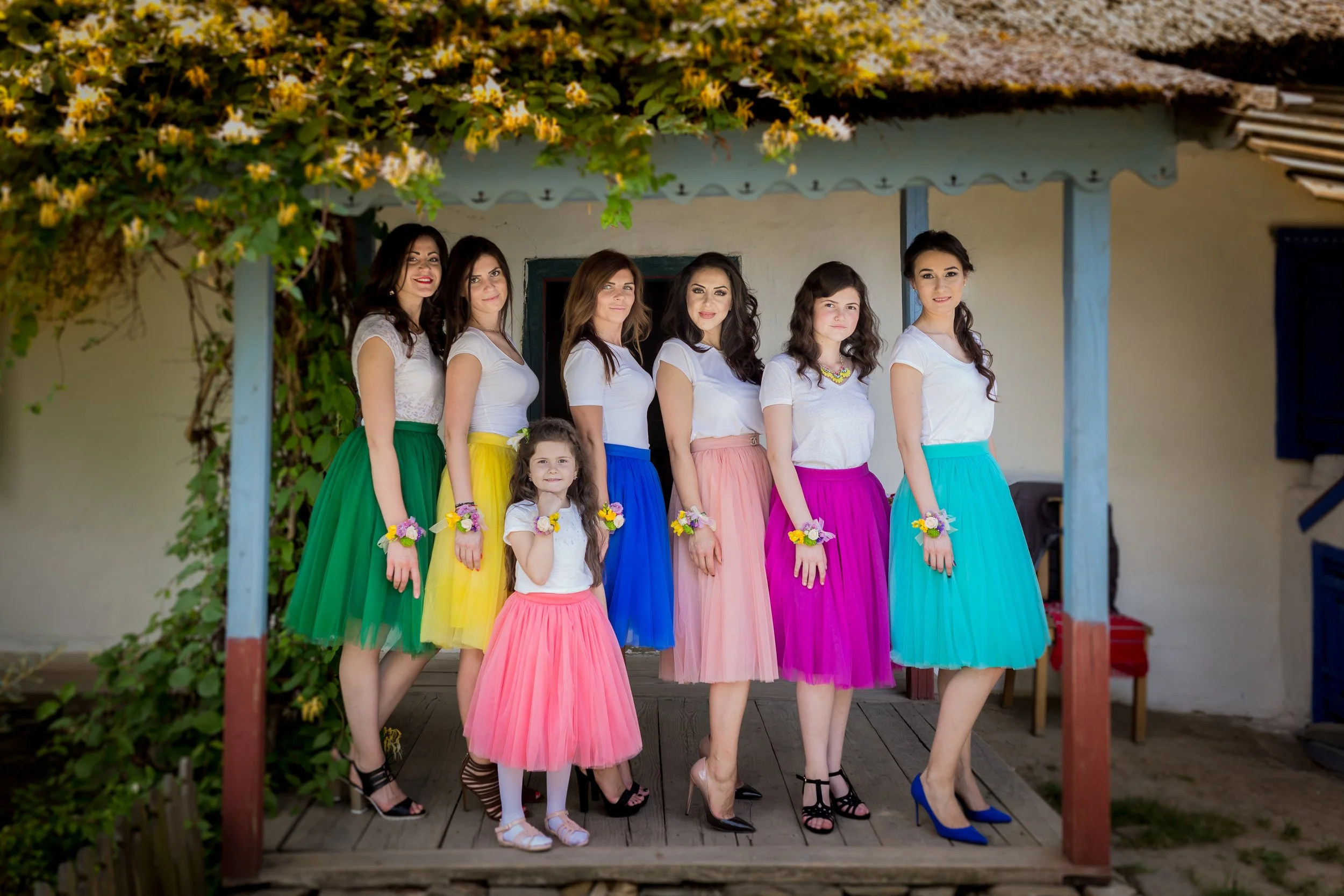 Six women and two girls standing on a porch, wearing colorful skirts and white tops, with some holding small bouquets of flowers, under a vine-covered structure.