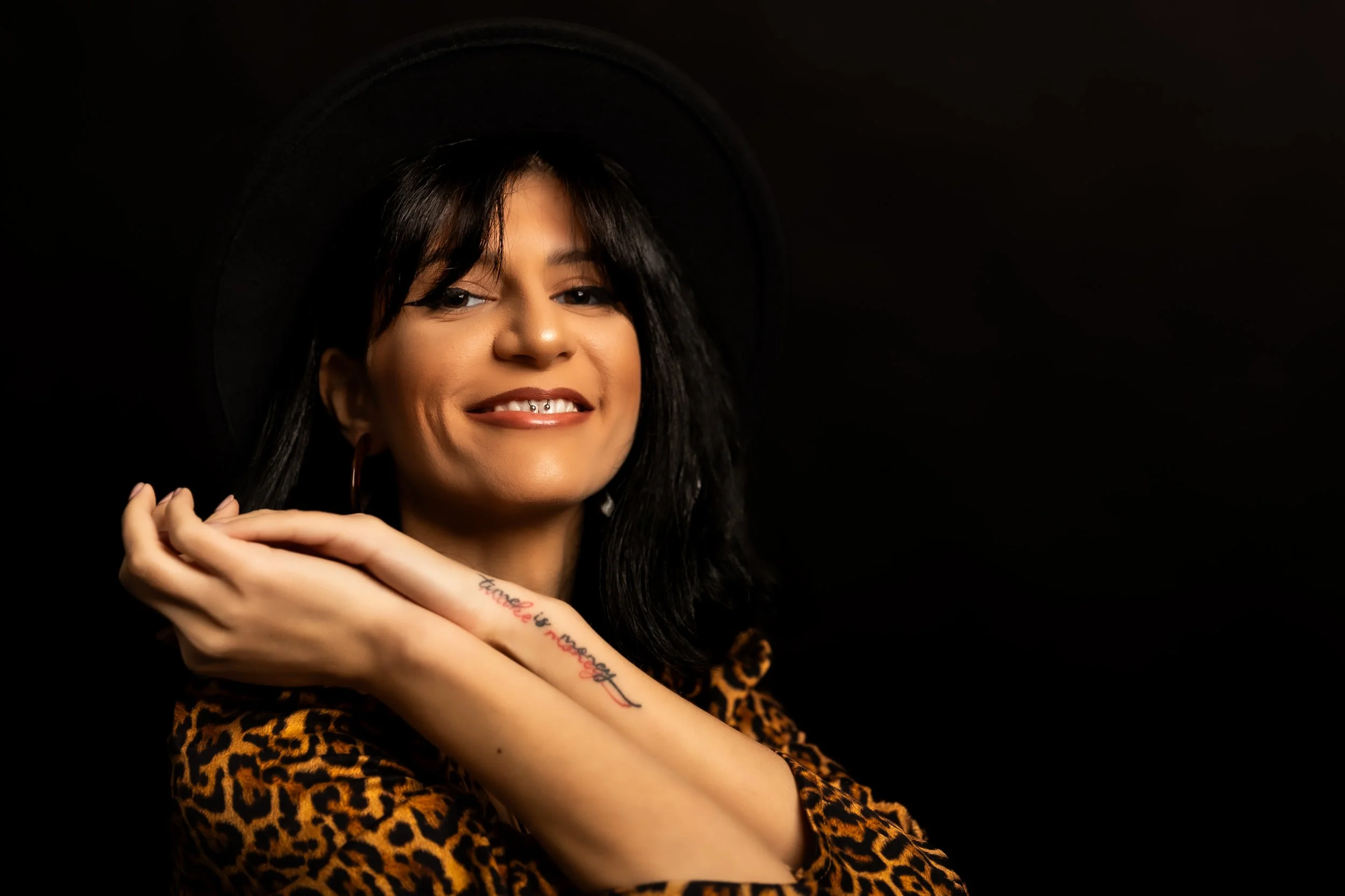 A woman with dark hair and a black hat smiling, wearing a leopard print top, with her hands crossed in front of her chest showing a tattoo on her left forearm that says 'Love is enough' in red and black ink, against a plain dark background.