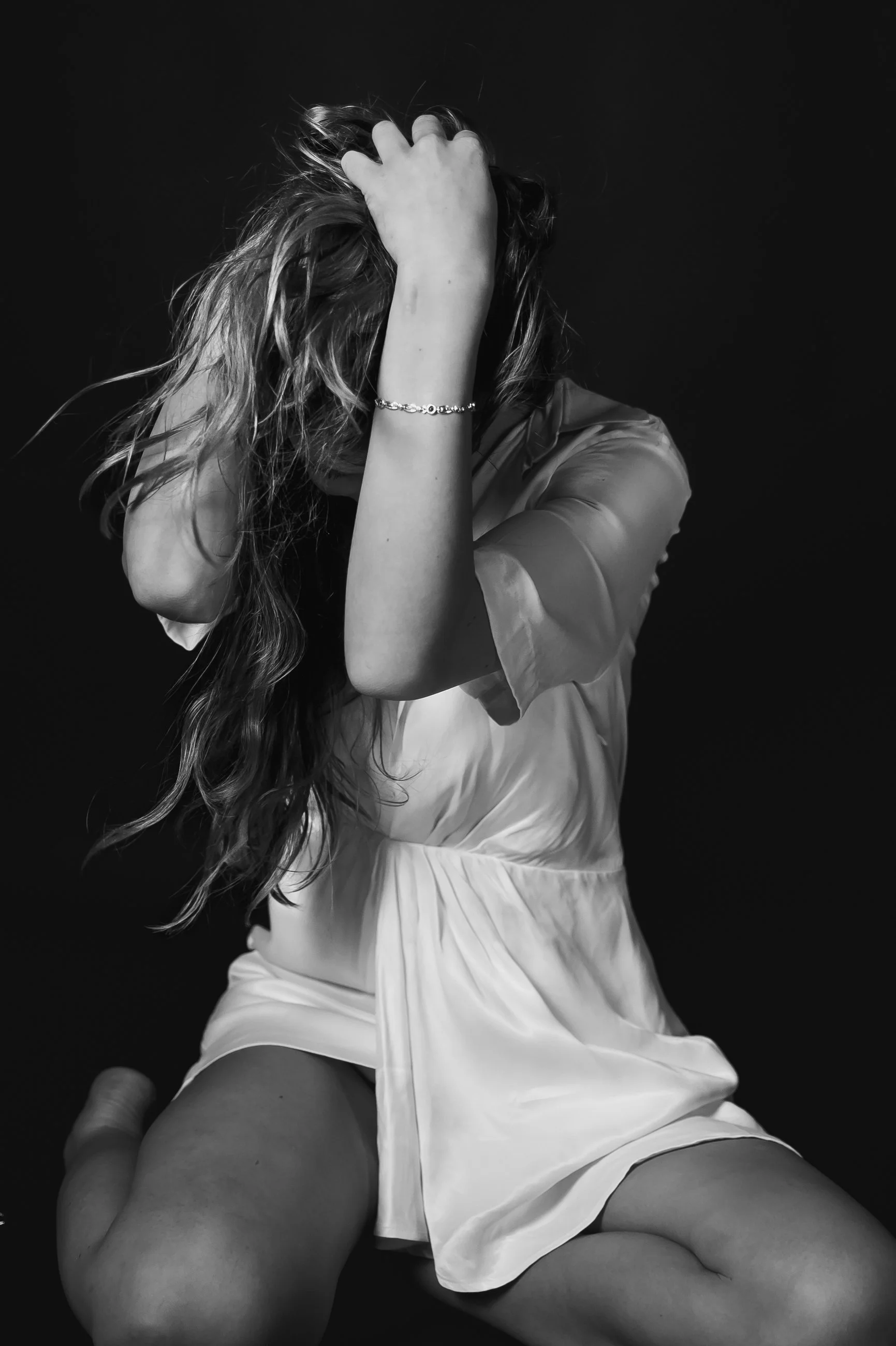 A woman with long, wavy hair, wearing a bracelet, resting her head and hand on her hair. She is dressed in satin clothing, sitting against a dark background.