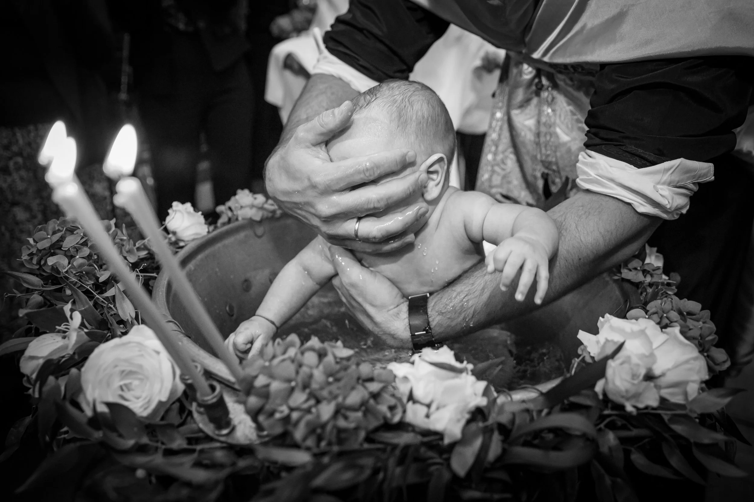 A person performing a baptism in a baptismal font, holding a baby under water with water splashing around, surrounded by flowers and candles.