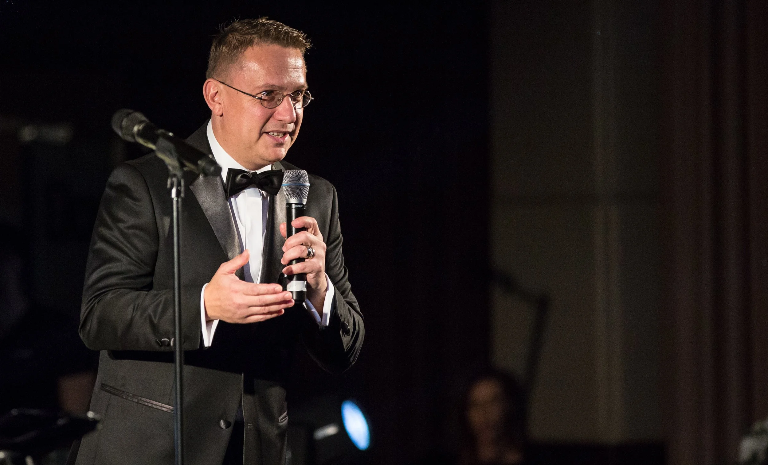 A man in a tuxedo with glasses speaks into a microphone on stage at an event.