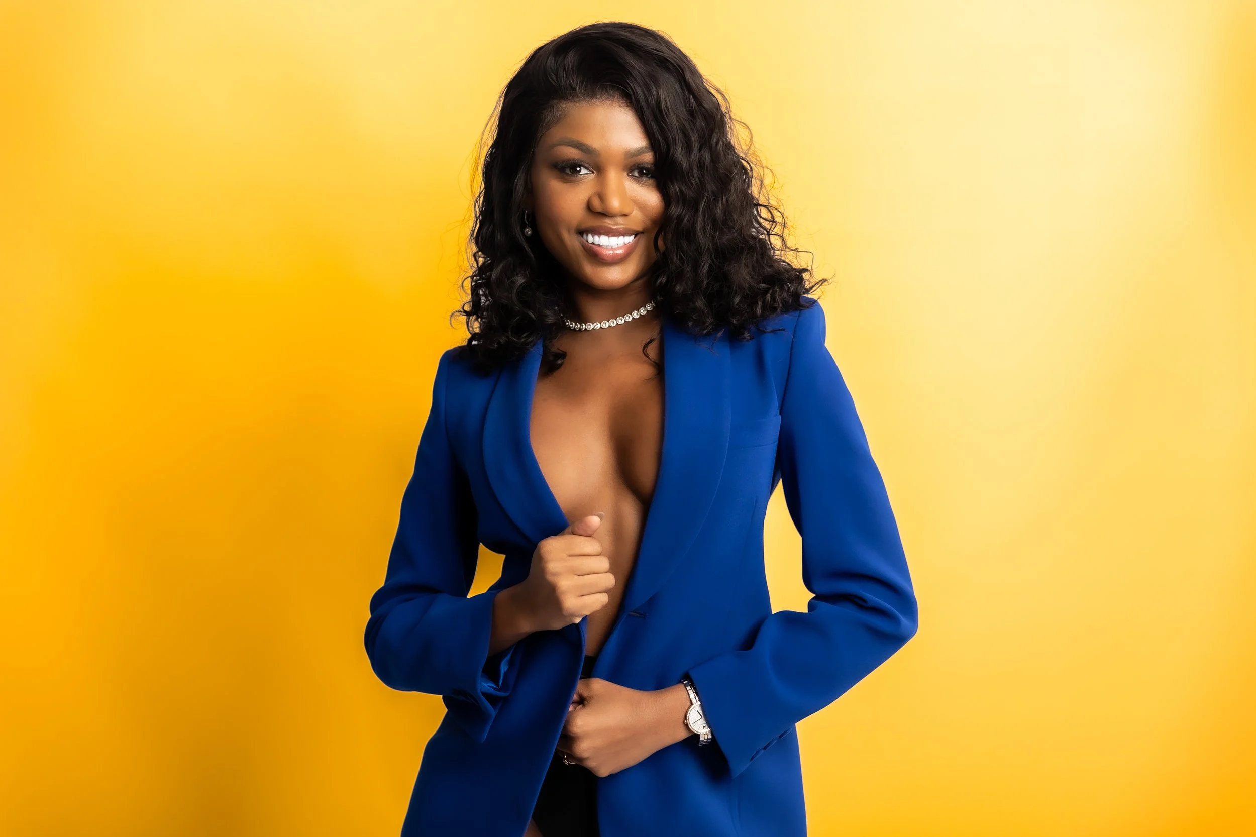 A woman with curly black hair wearing a blue blazer, pearl necklace, and watch, smiling in front of a yellow background.