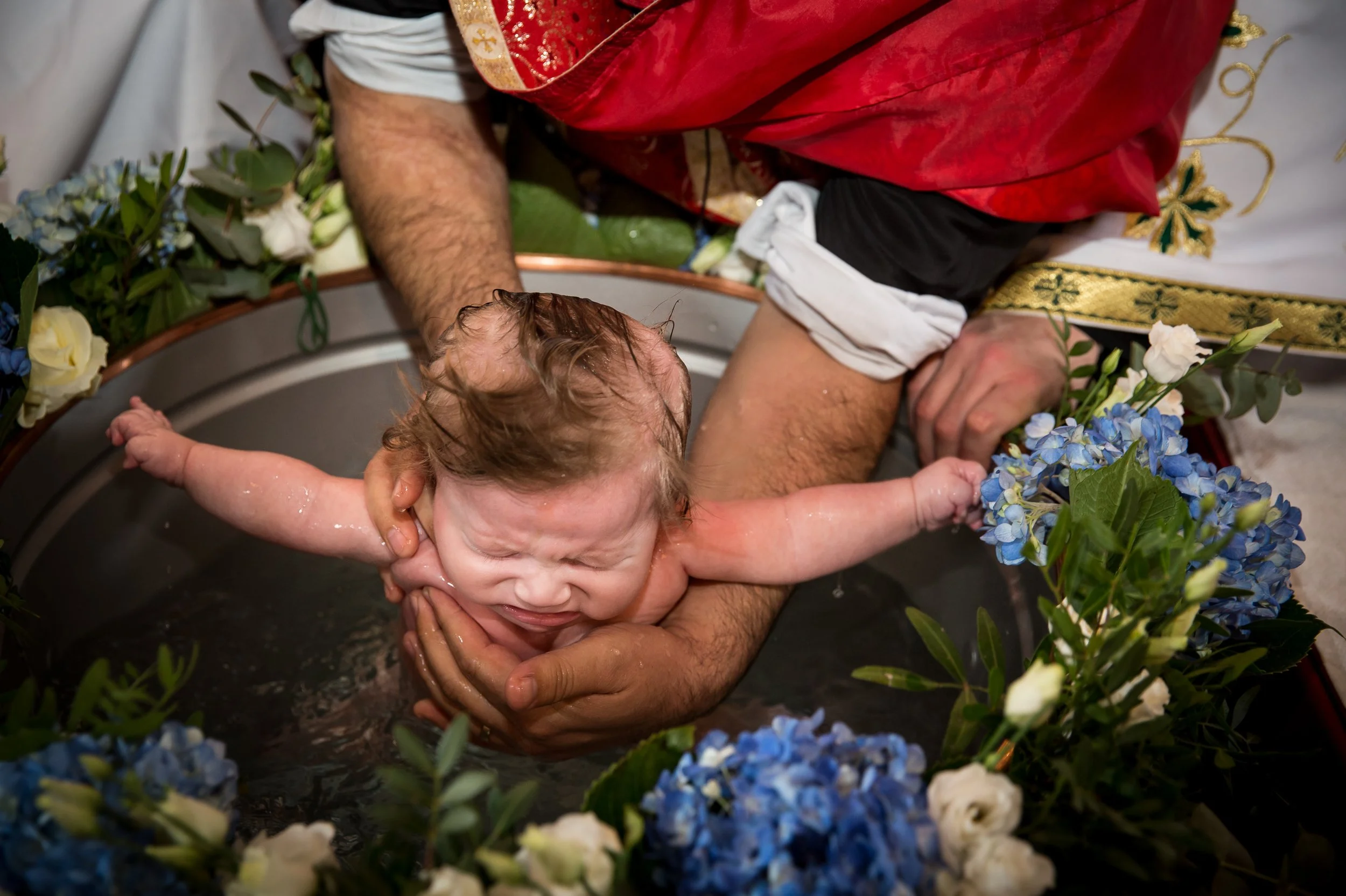 A baby being baptized in a ceremonial bath surrounded by blue and white flowers, with water on the baby's face and a person's arms supporting the baby.