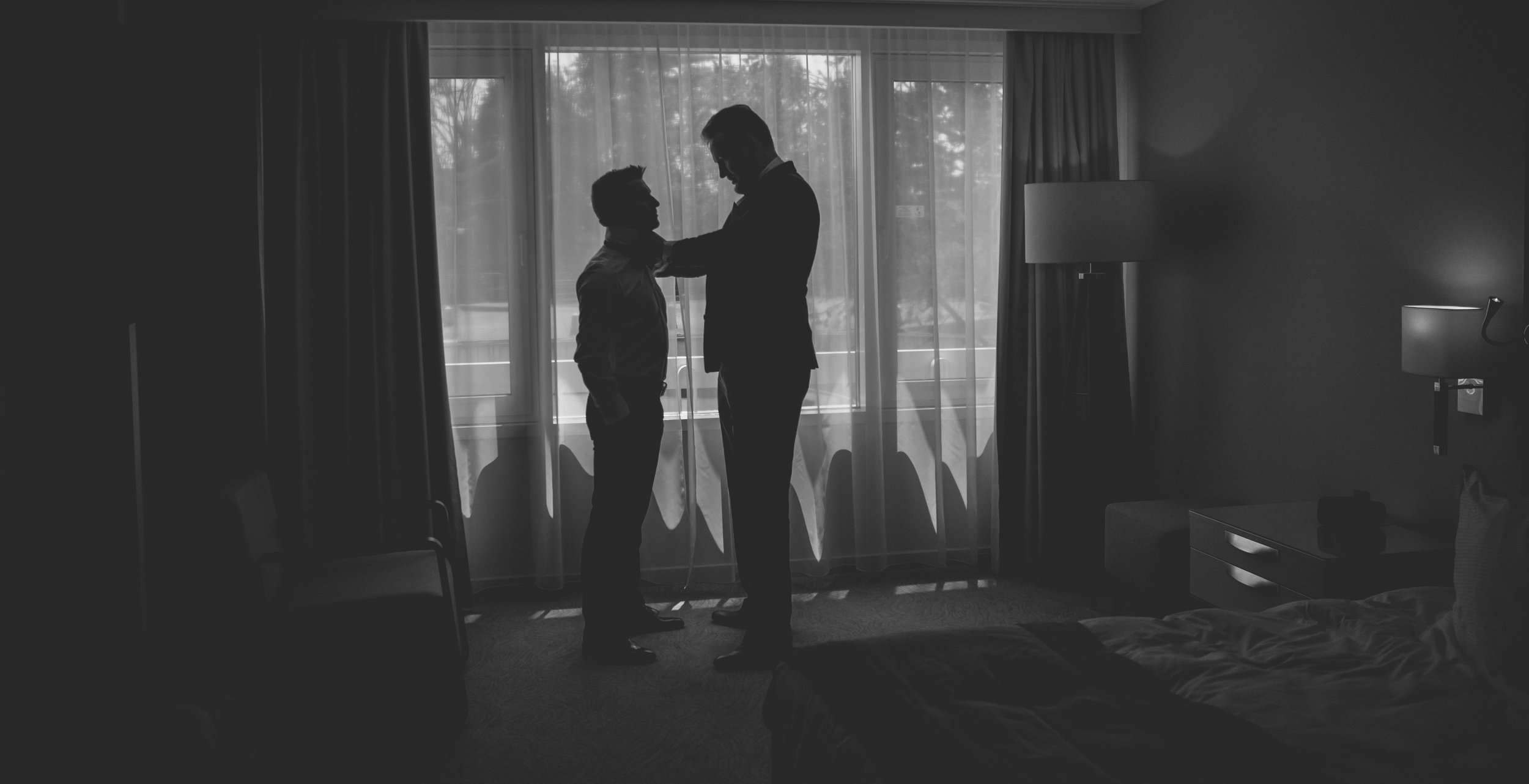 Silhouettes of two men in formal attire facing each other, standing in front of large windows with curtains, in a hotel room or apartment, during the daytime.