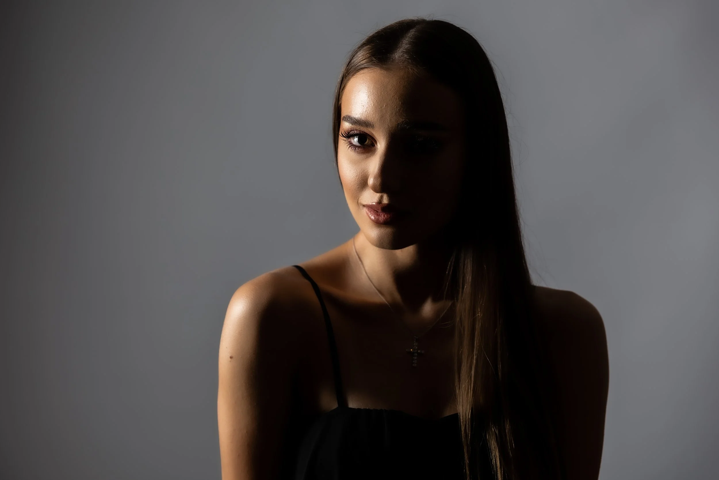 A woman with long brown hair, wearing a black spaghetti strap top, looking slightly to the side with one eye visible, against a plain gray background.