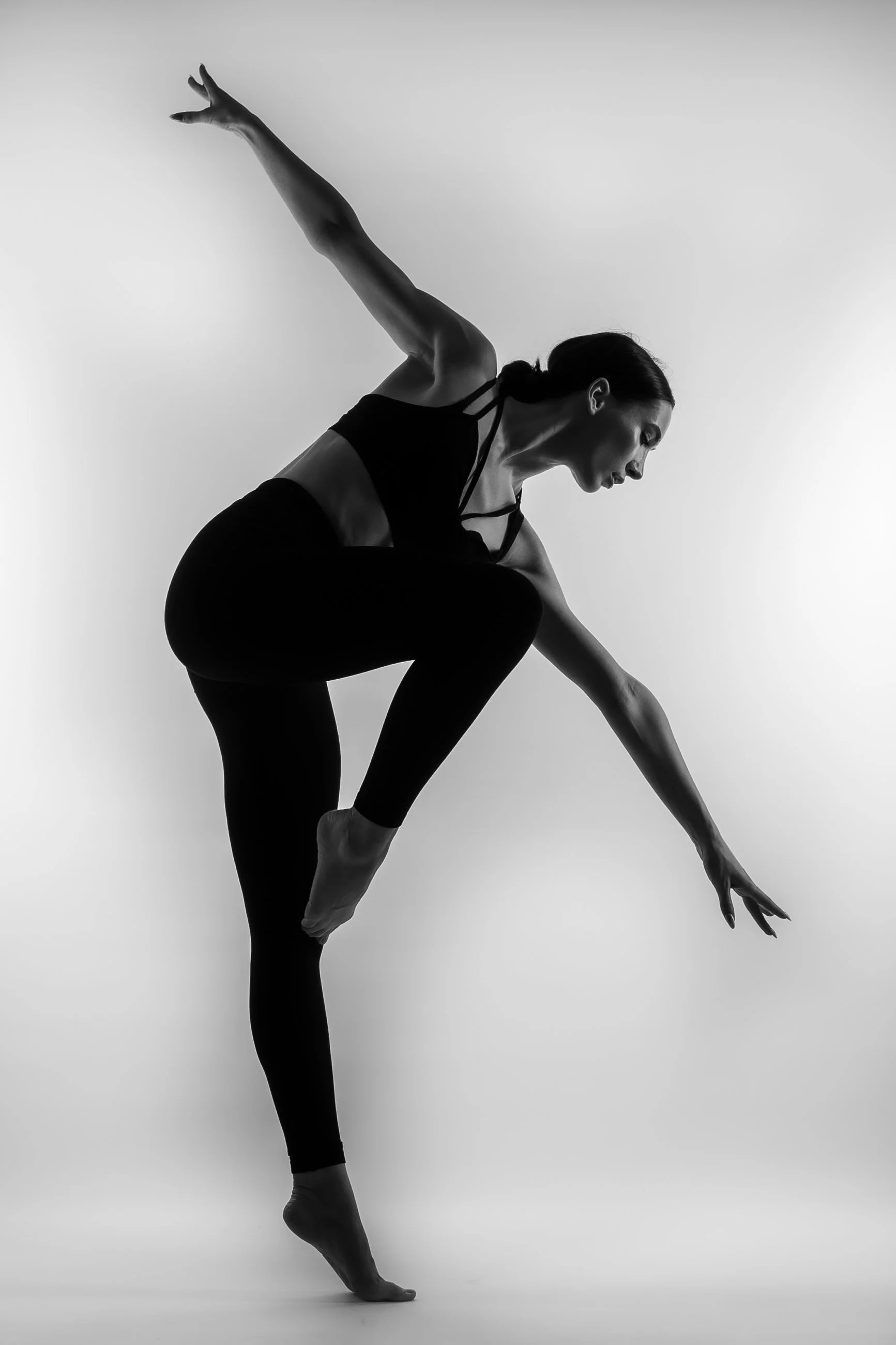 A black-and-white photo of a woman in a dance or yoga pose, balancing on one leg with her other leg bent and raised behind her. She is reaching forward with one arm, while the other arm extends back, and she is wearing a sports bra and leggings.