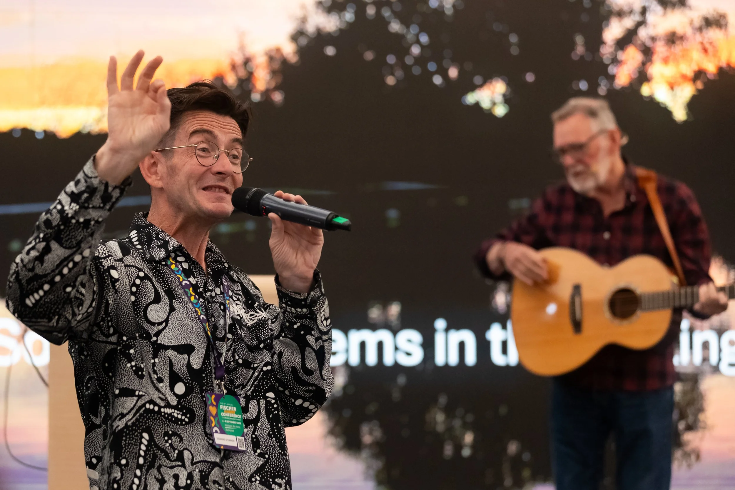 A man with glasses and a patterned shirt holding a microphone and raising his hand, with a woman playing guitar in the background, at an indoor event.