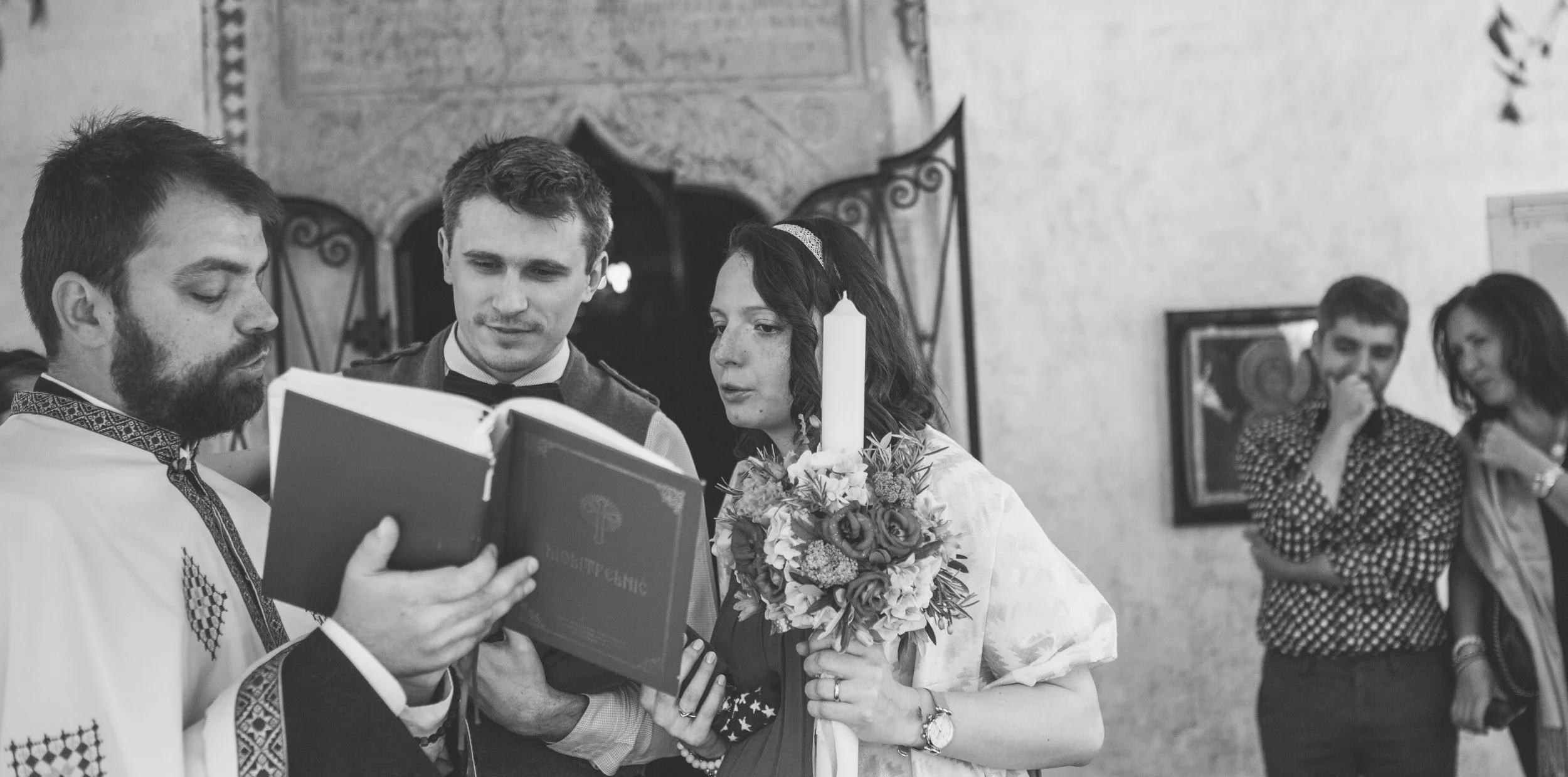 A couple in wedding attire standing with a priest during a wedding ceremony, holding a bouquet and looking at a book, with two women in the background.