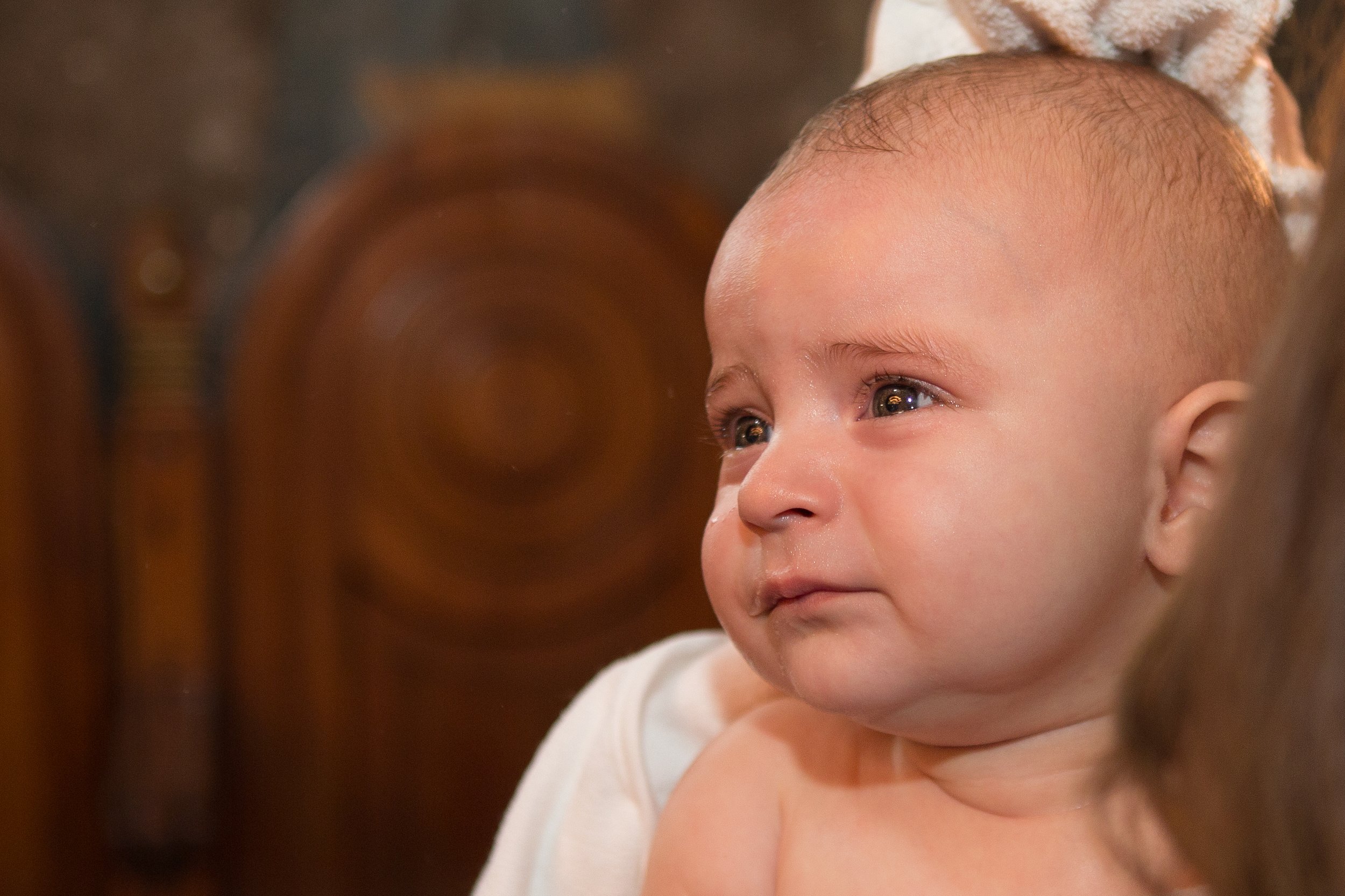 Close-up of a baby with brown eyes and light skin, wearing a white outfit, lying on a surface with a wooden and blurred background.